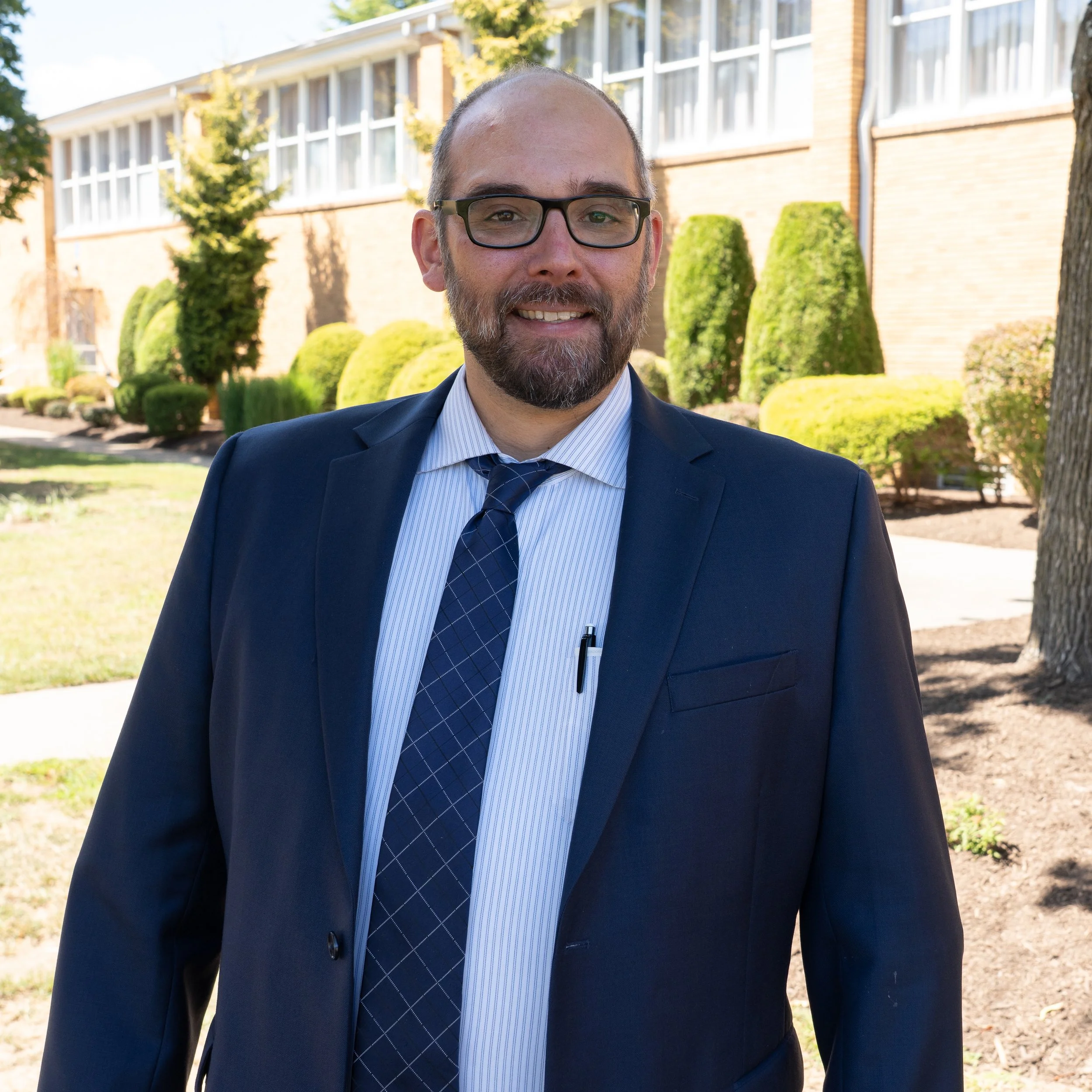 A man with glasses and a beard wearing a blue suit, white checked shirt, and a navy polka dot tie, smiling against a gray background.