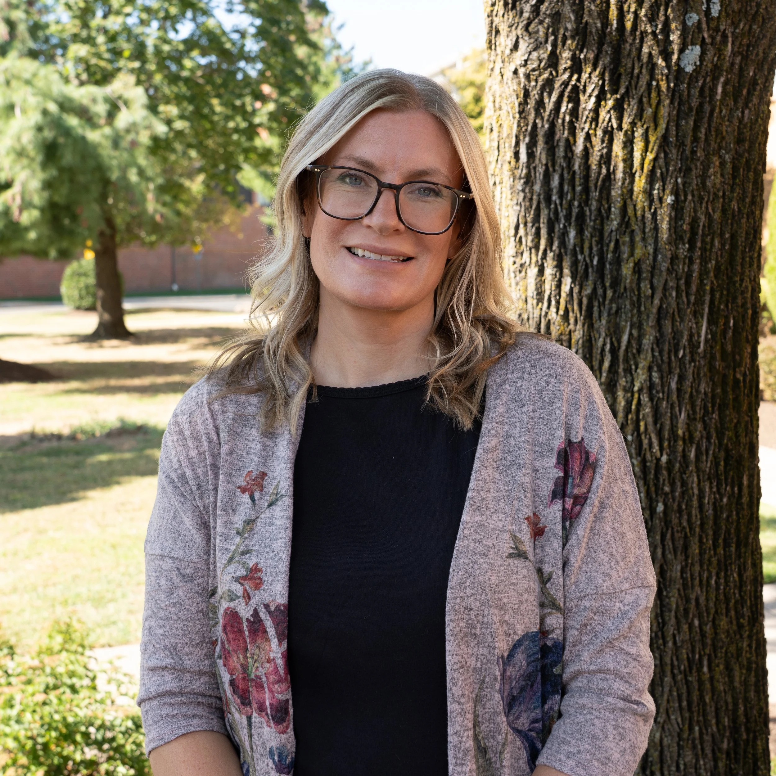 Woman with long blonde hair, wearing glasses and a small beige top, smiling at the camera against a grey background.
