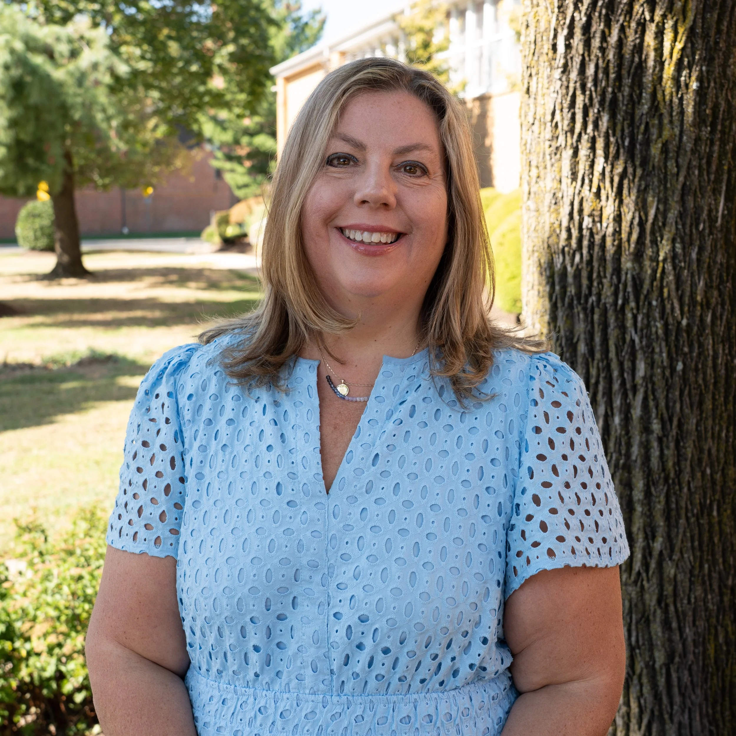 A smiling woman with shoulder-length wavy blonde hair, wearing a coral-colored top with a decorative pattern and a small pendant necklace, posed against a gray background.
