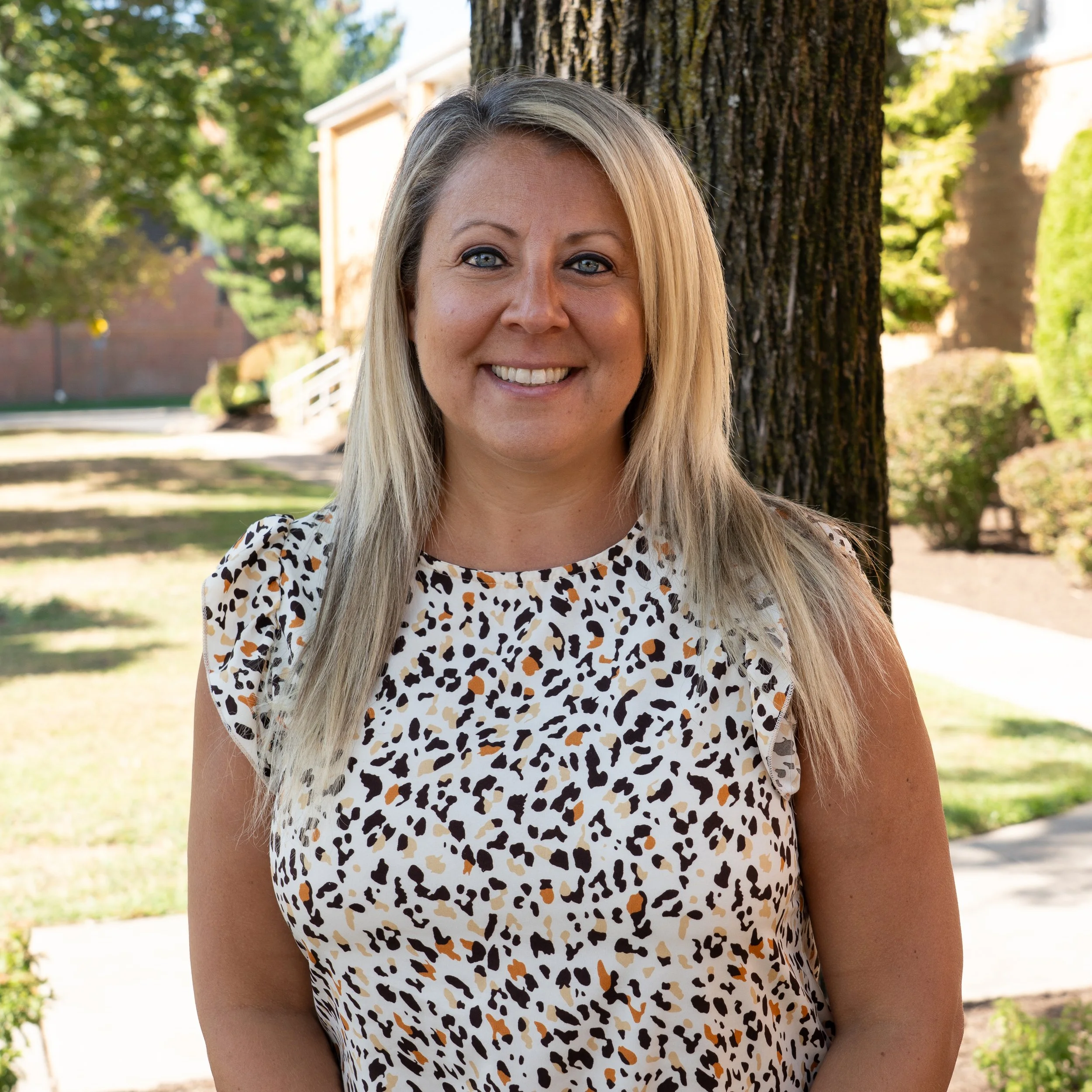 A woman with blonde hair in a ponytail, smiling, wearing a navy blue floral blouse, against a gray backdrop.