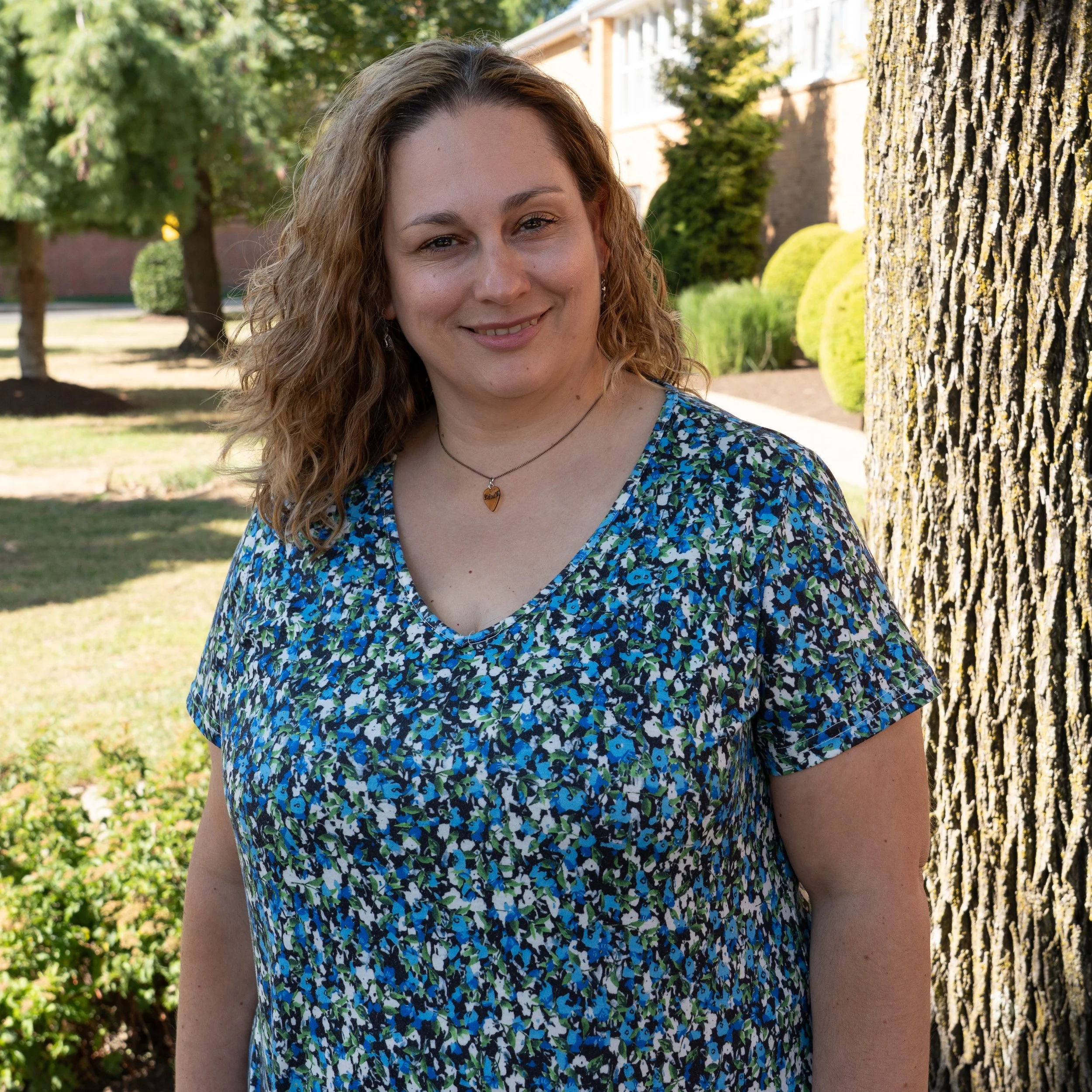 A woman with curly brown hair tied back, wearing hoop earrings, a white top with black accents, and a pink necklace.