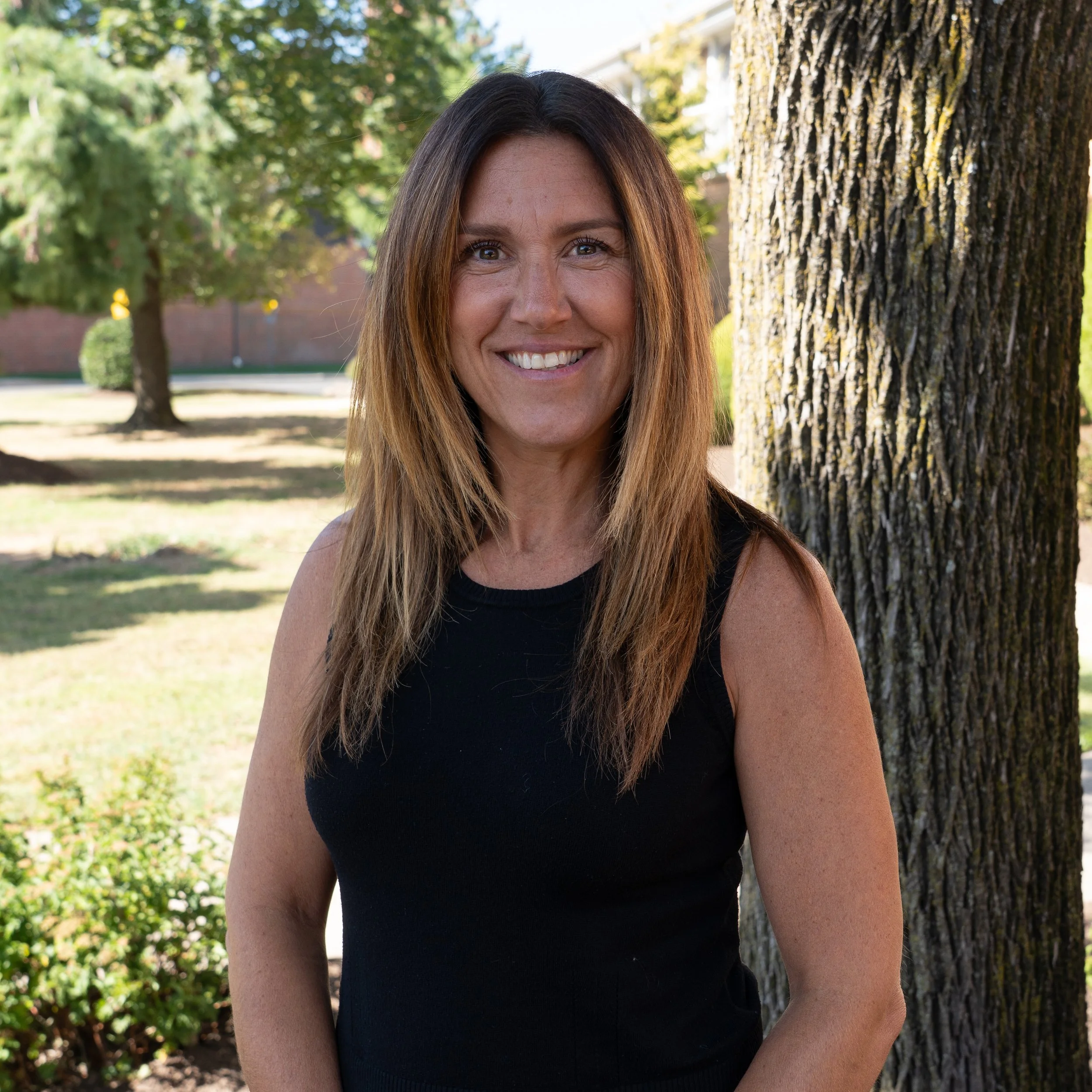 Portrait of a woman with straight, shoulder-length brown hair, smiling, wearing a white top, against a gray background.