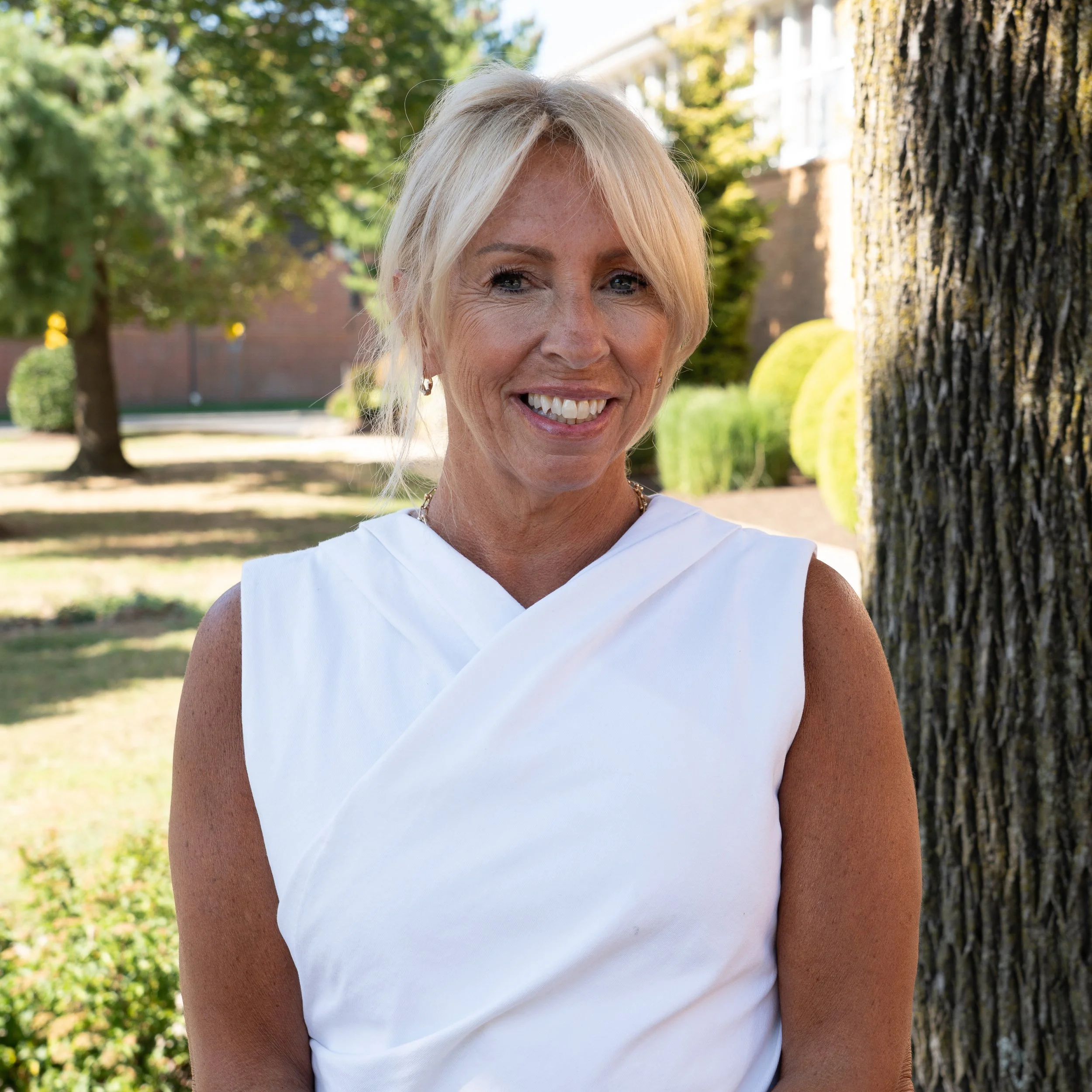 A woman with blonde hair, blue eyes, wearing a white top, and a gold necklace, posing against a gray background.