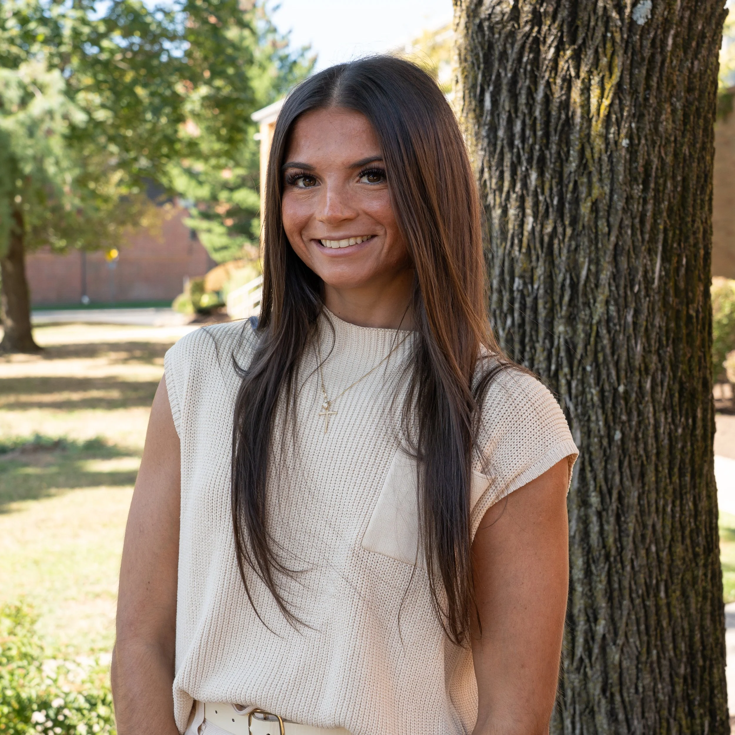 Woman with long dark hair smiling, wearing a pink shirt and layered necklaces, against a gray background.