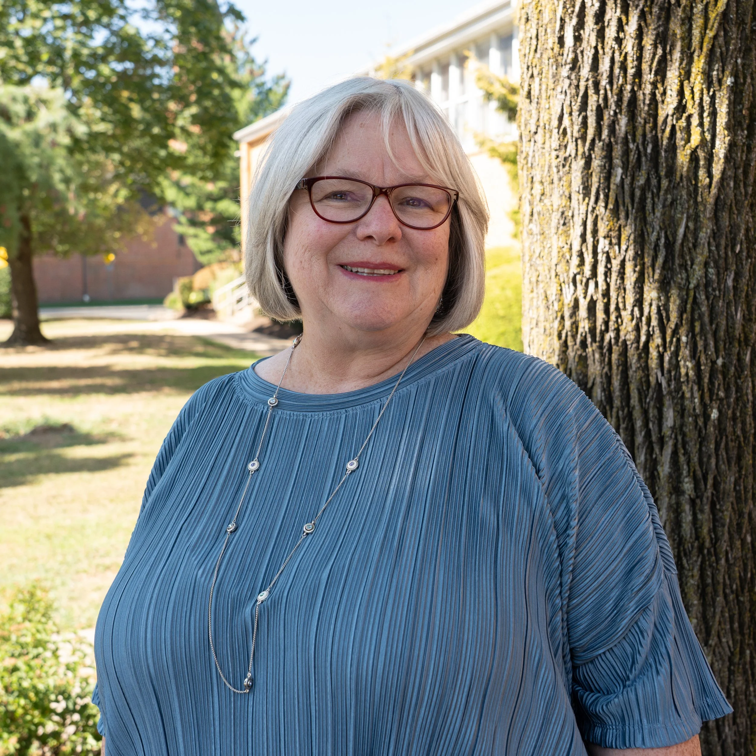 A middle-aged woman with long, wavy brown hair wearing glasses and a pink top. She has a black lanyard around her neck with gold writing and a grey background.