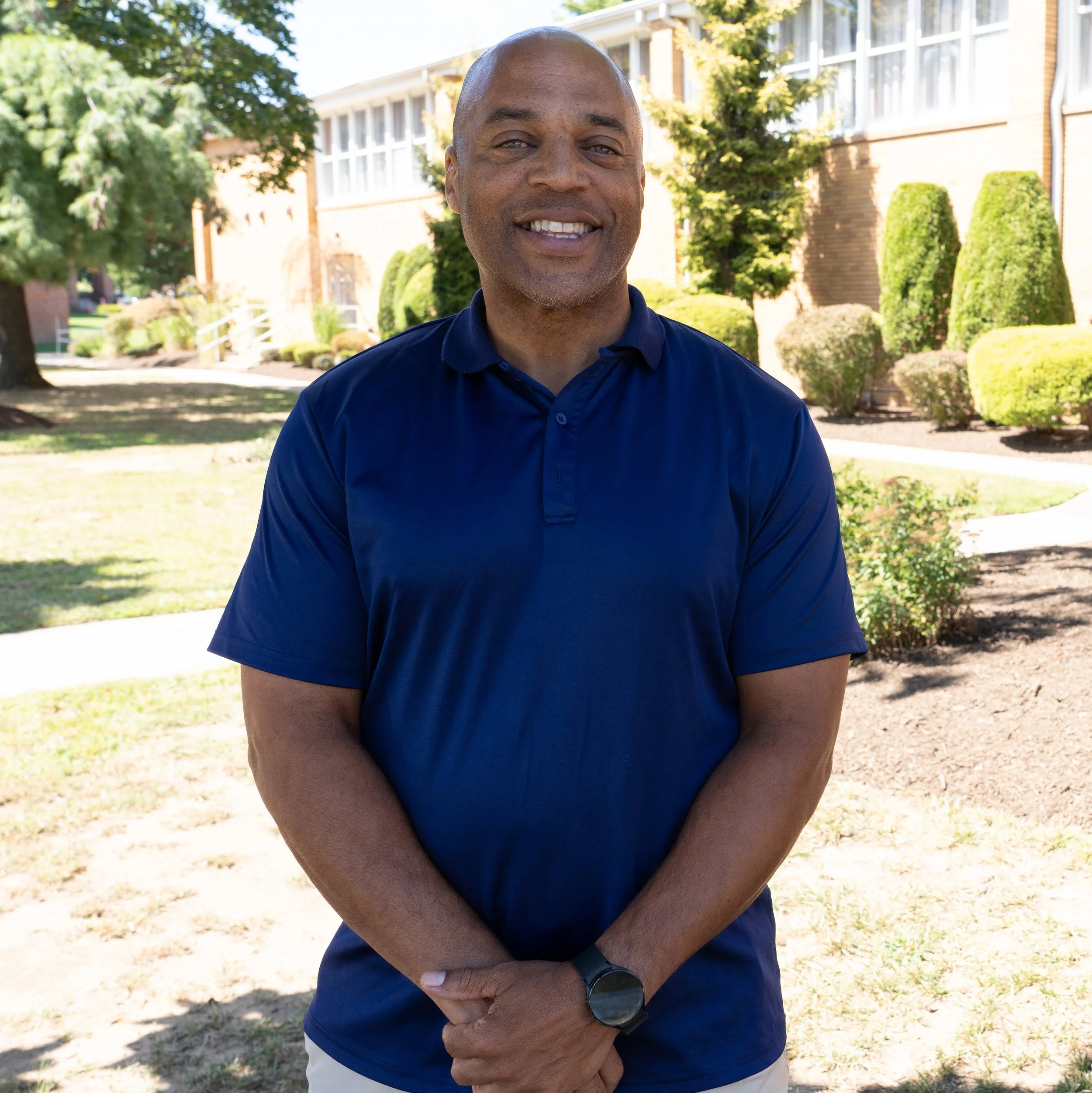 A smiling middle-aged man with a shaved head, gray beard, and blue eyes, wearing a pink collared shirt, photo portrait against a dark gray background.