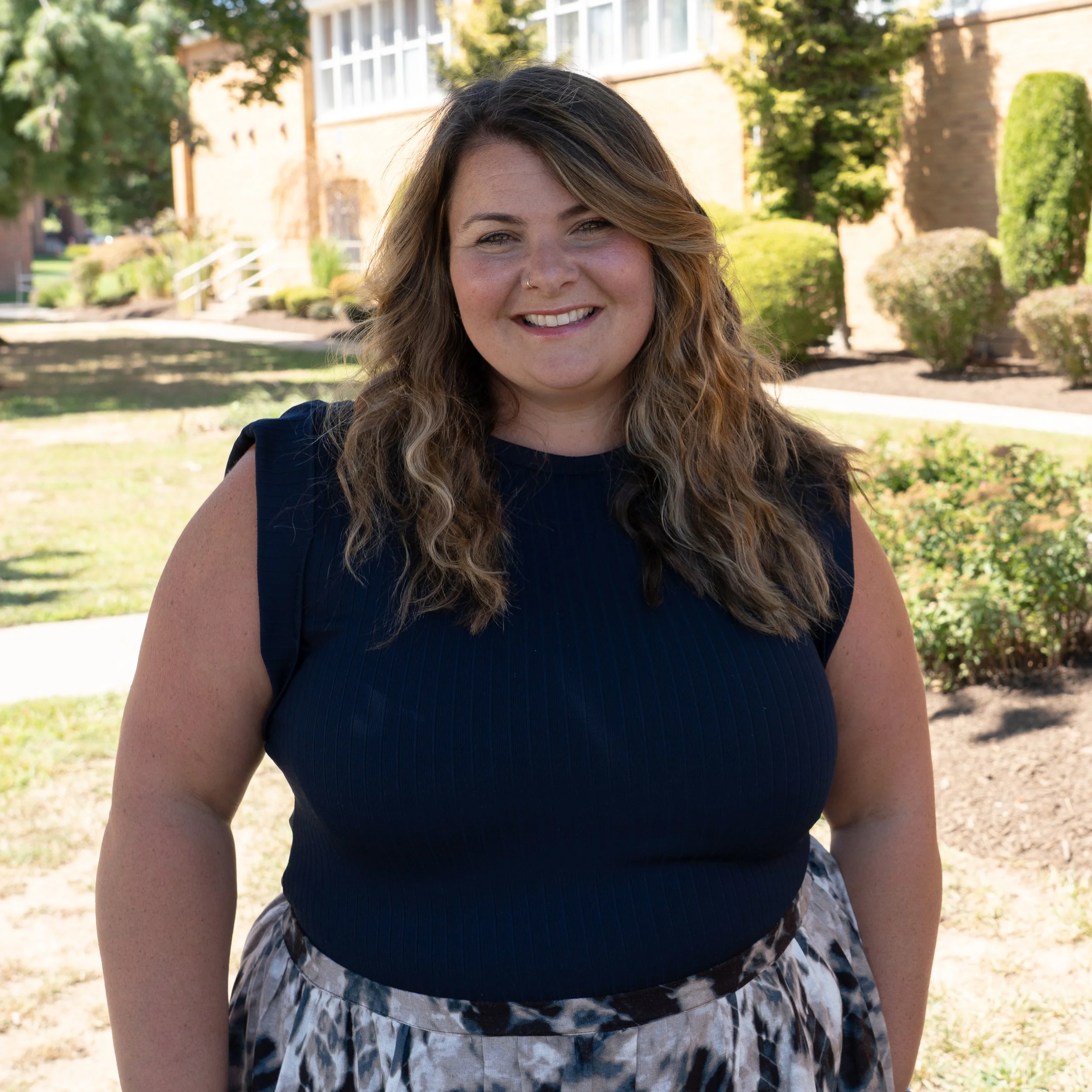 Headshot of a woman with long, wavy brown hair and a black top, smiling at the camera against a gray background.