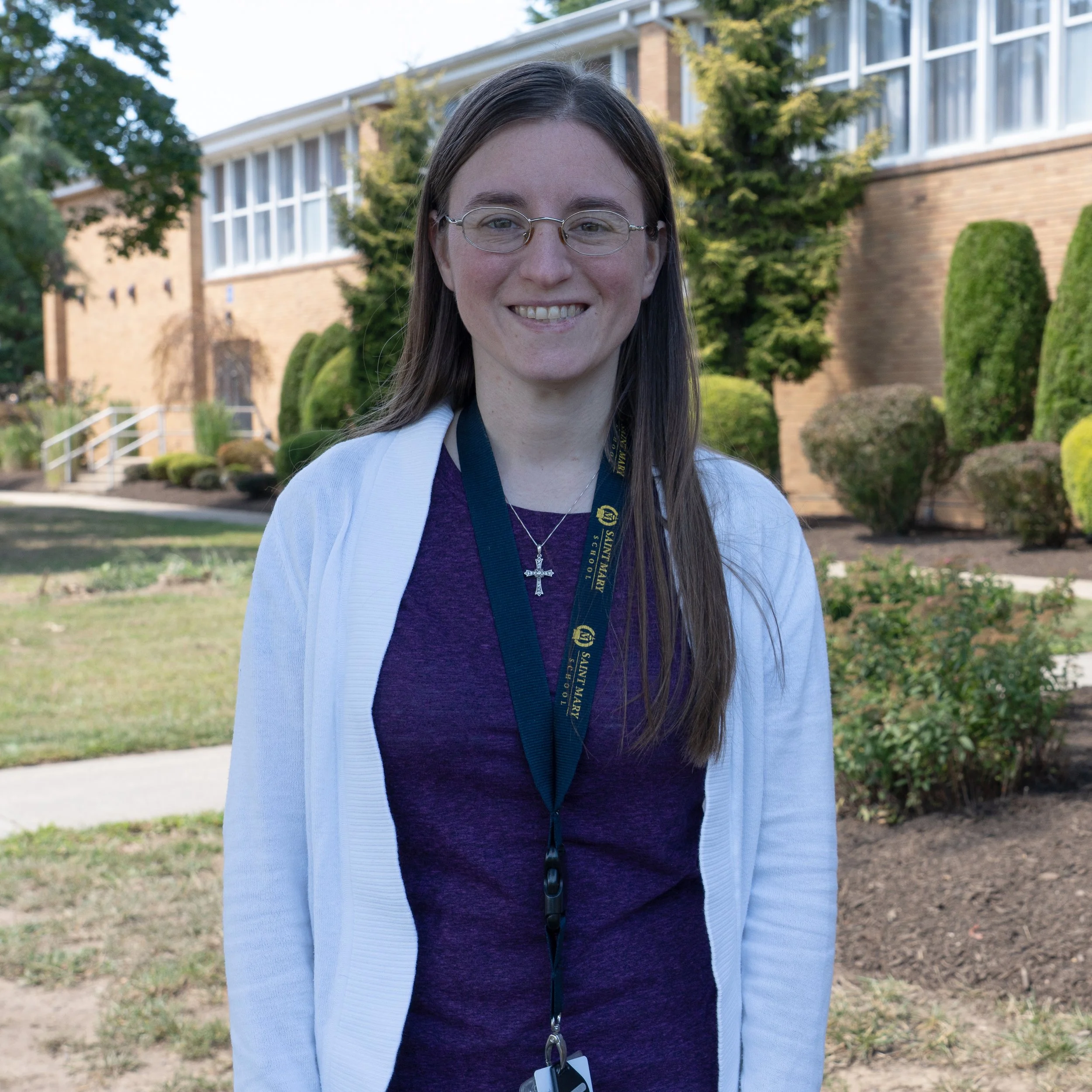 A woman with long brown hair, glasses, and a friendly smile wearing a green shirt and gray hoodie against a gray background.