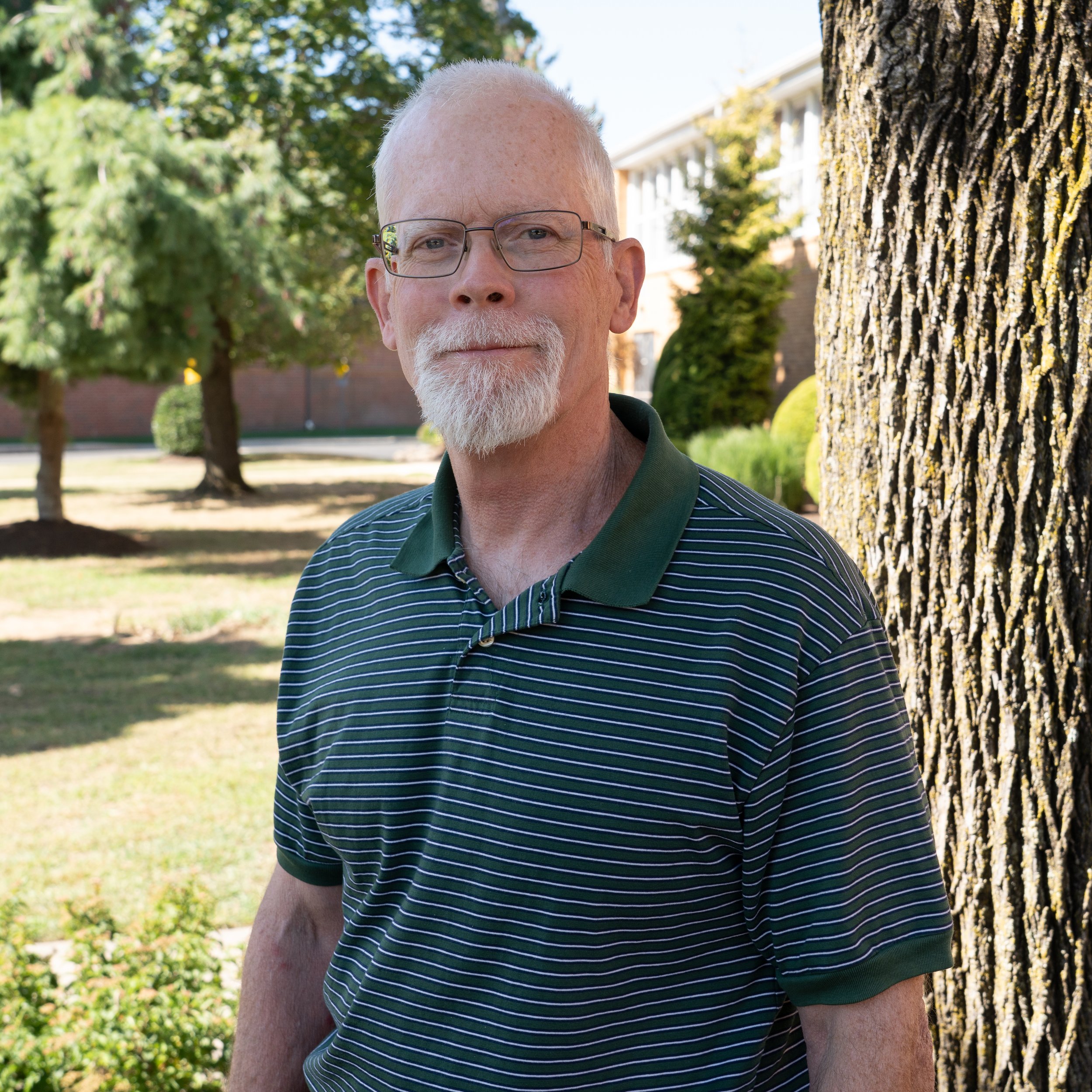 Portrait of an elderly man with glasses, a white beard and mustache, wearing a green and white striped collared shirt, against a gray background.