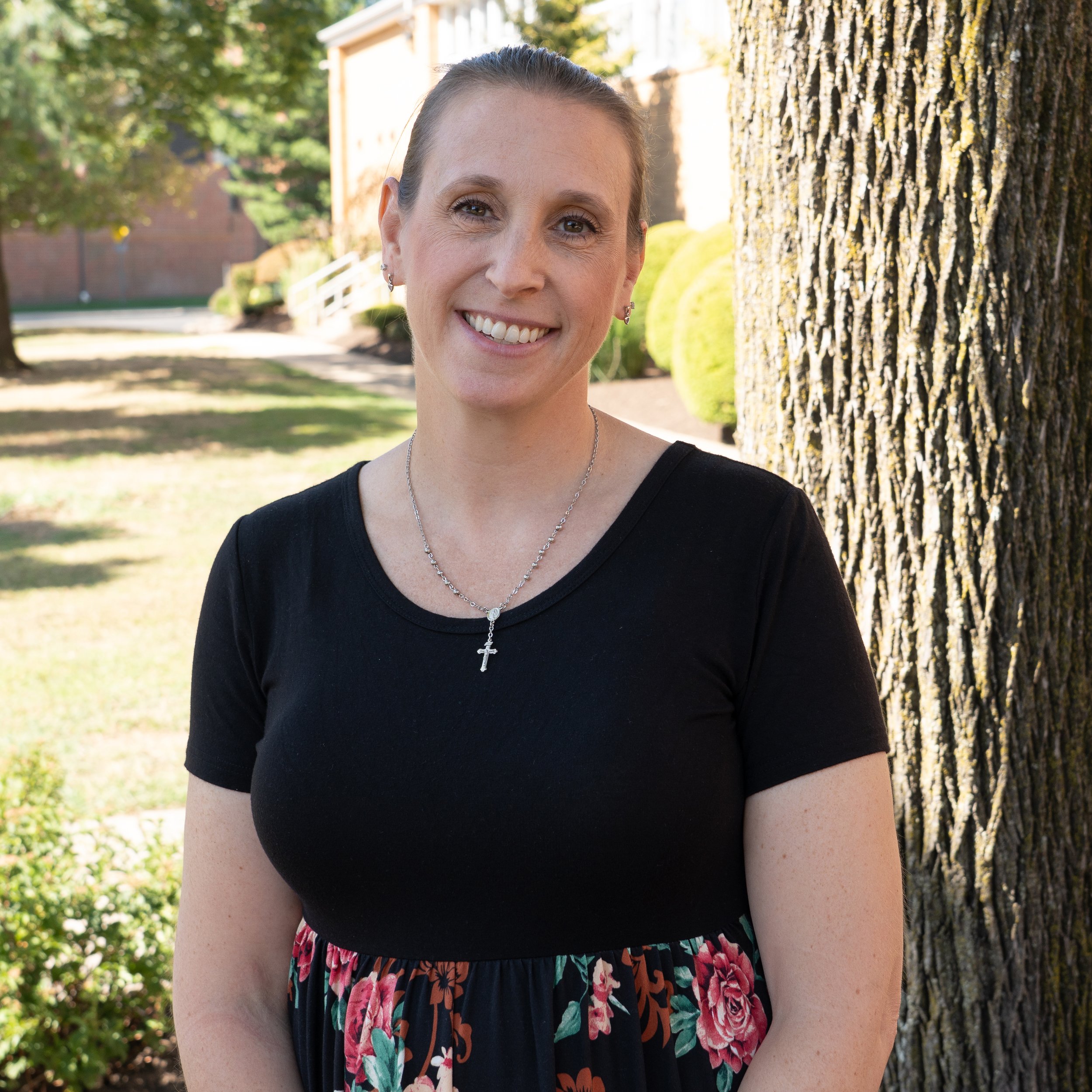 A woman with light skin, brown hair tied back, wearing a black top, a necklace with a cross pendant, and earrings, smiling at the camera with a gray background.