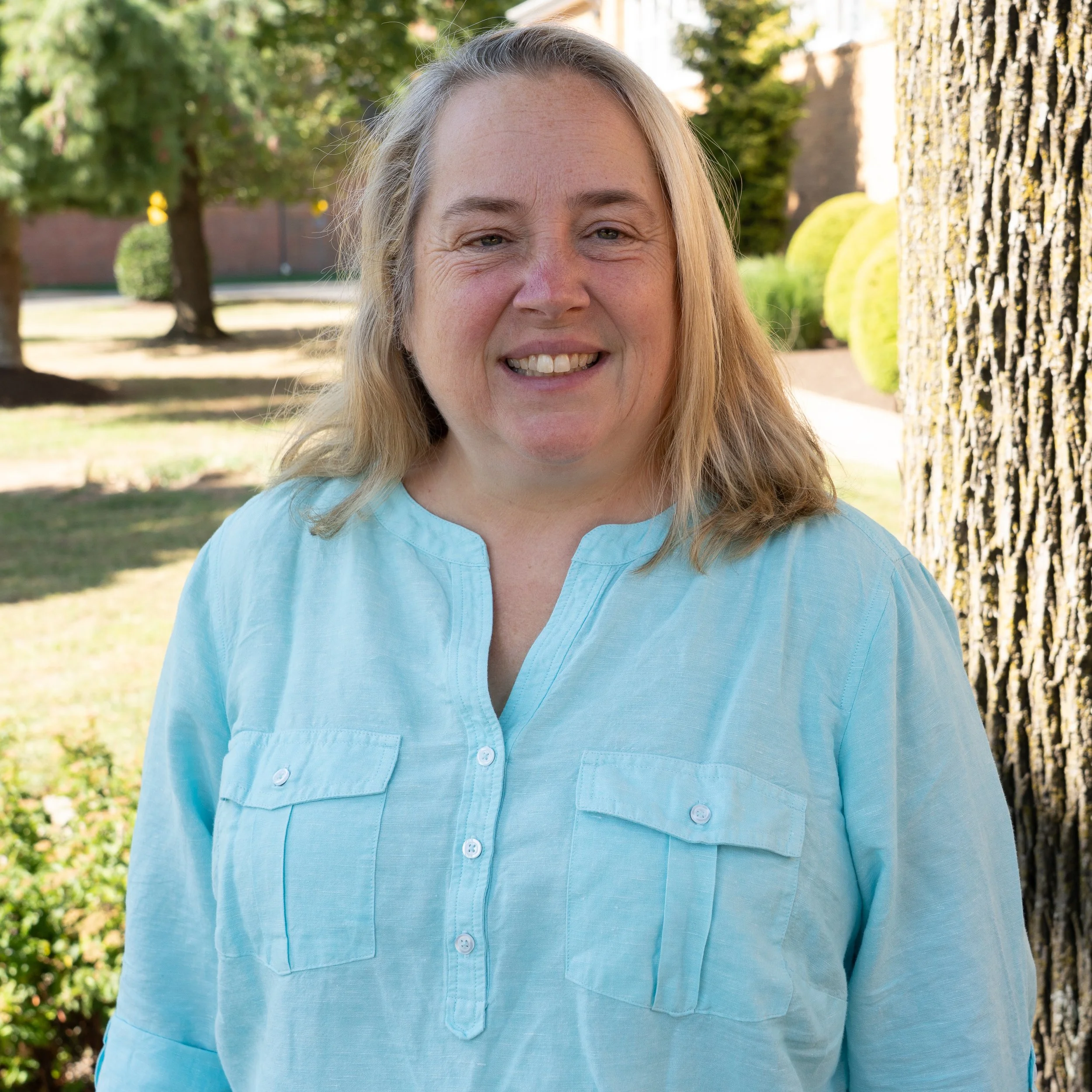 A smiling woman with long blonde hair wearing a pink shirt against a gray background.