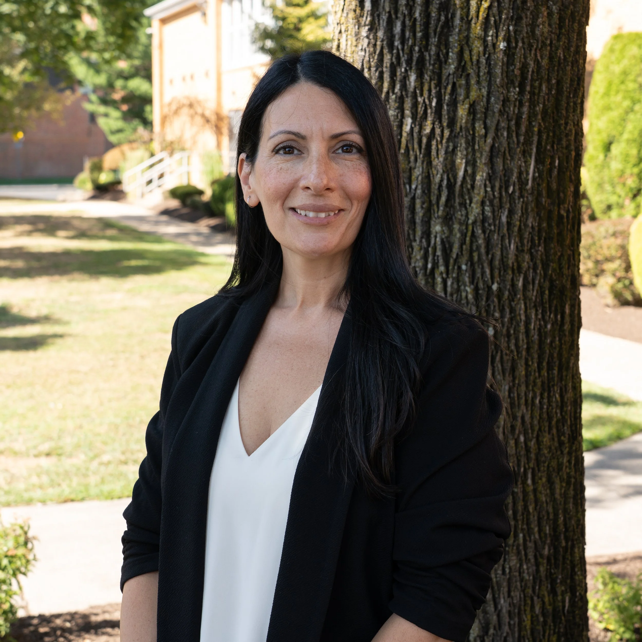 A woman with long black hair, light skin, smiling, wearing earrings and a black top, against a gray background.