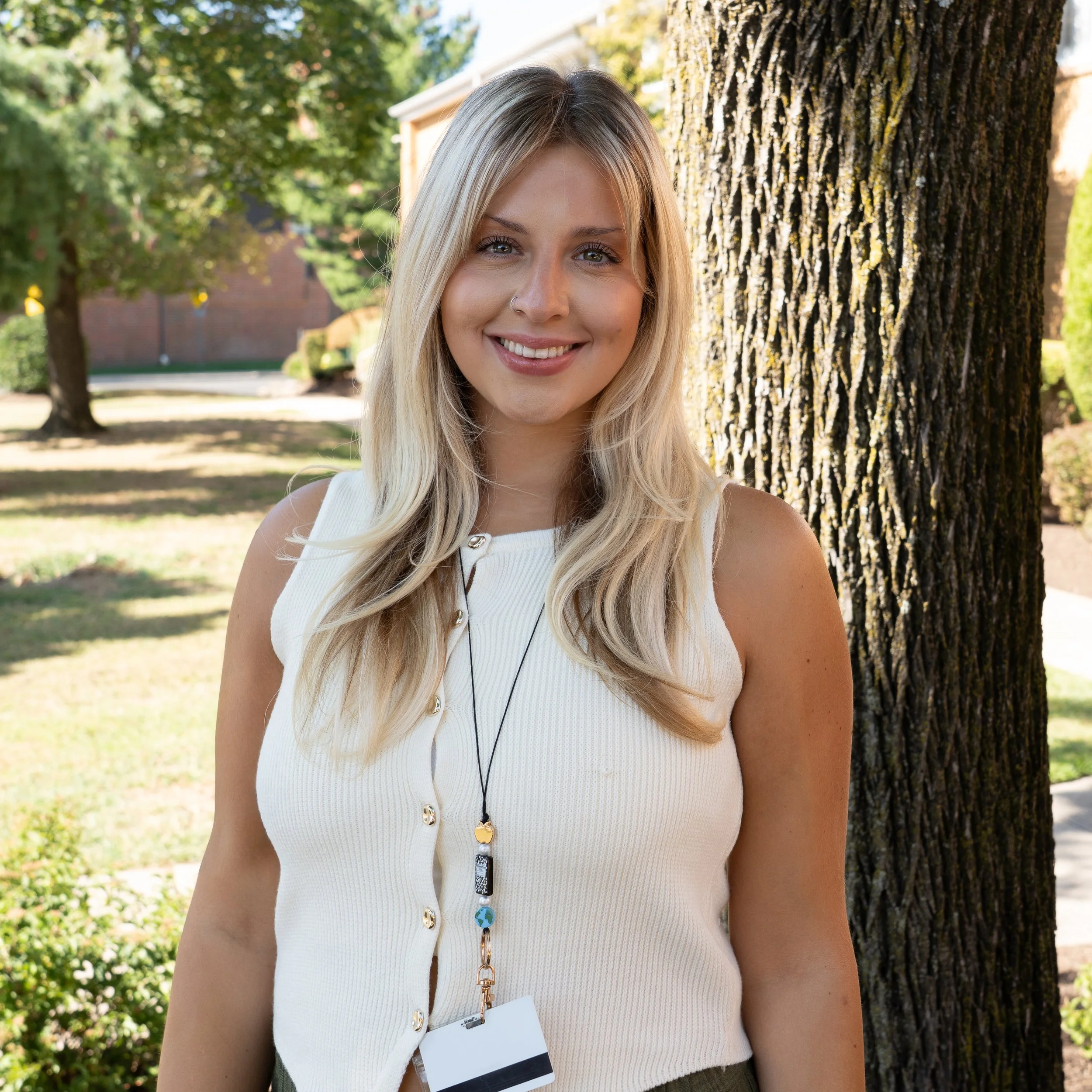 A young woman with blonde hair, wearing a navy sleeveless top, a cross necklace, and a lanyard with 'Summit Academy' printed on it, smiling against a grey background.