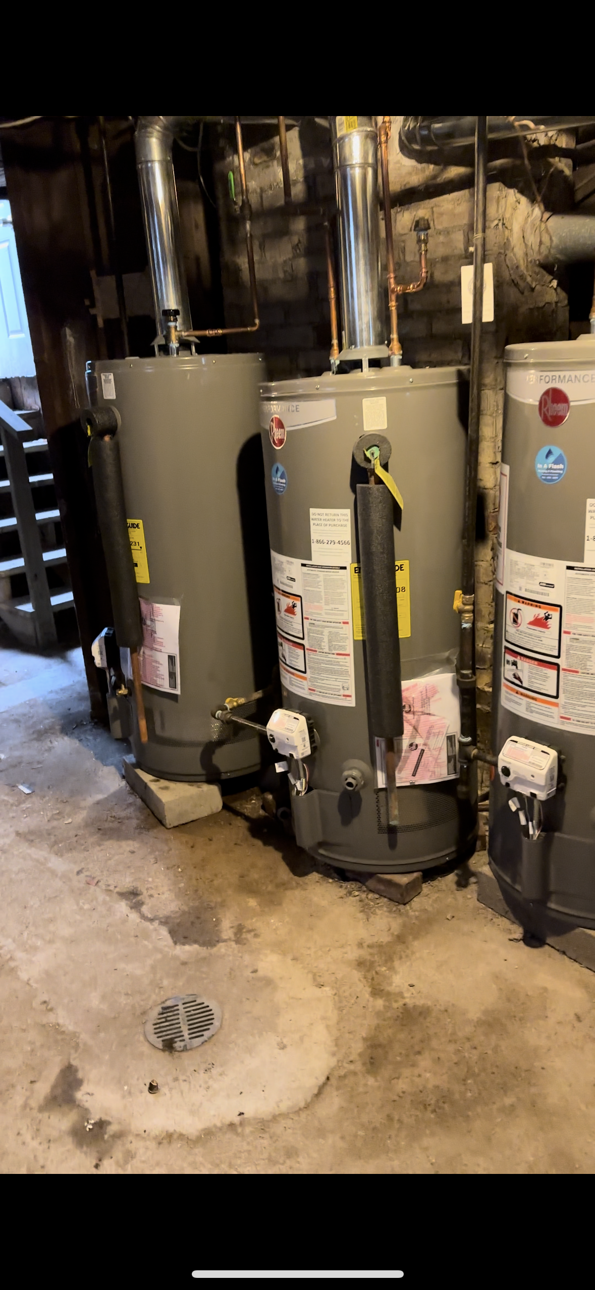 Three water heaters in a basement with pipes and a floor drain.
