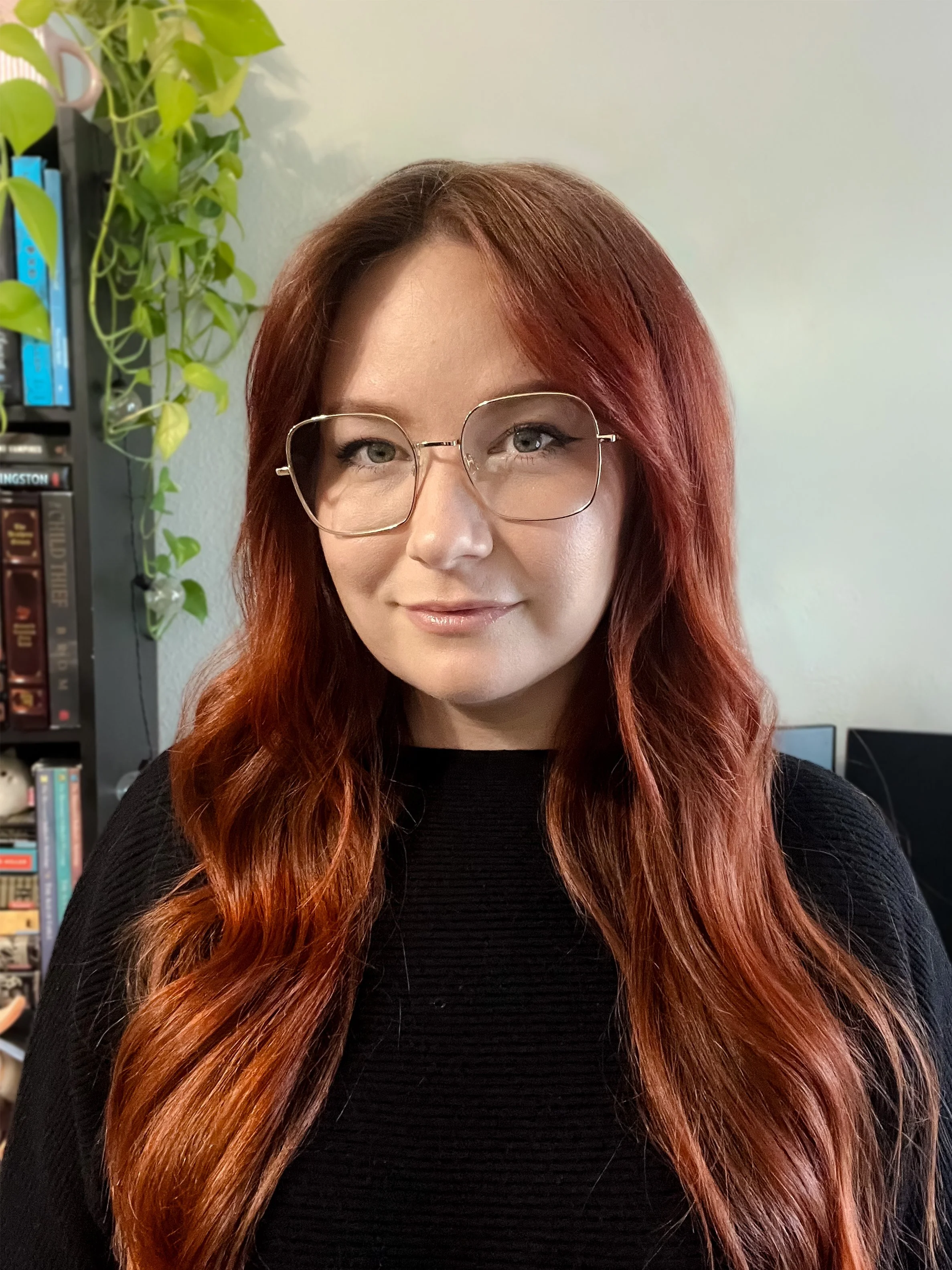 A woman with long wavy red hair and glasses poses for a photo indoors, with a bookshelf and some greenery in the background.