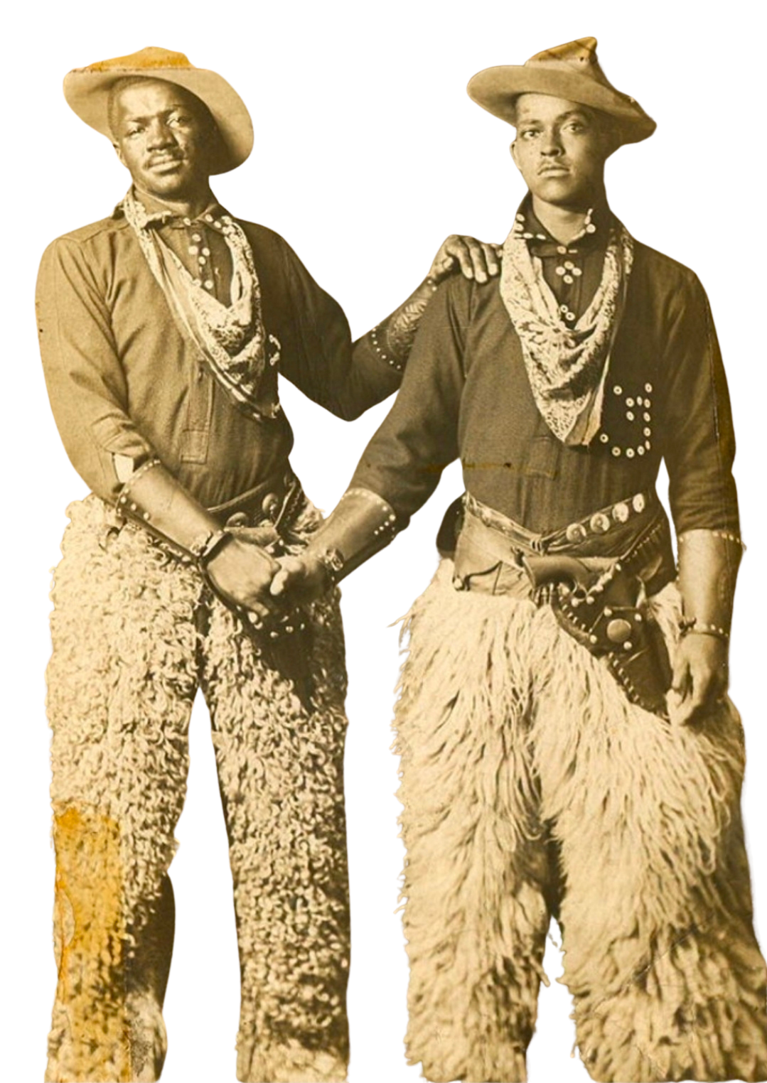 Two men dressed in old-fashioned cowboy outfits, with wide-brimmed hats, bandanas, and shaggy chaps, standing close together holding hands, one man has a hand on the other's shoulder.