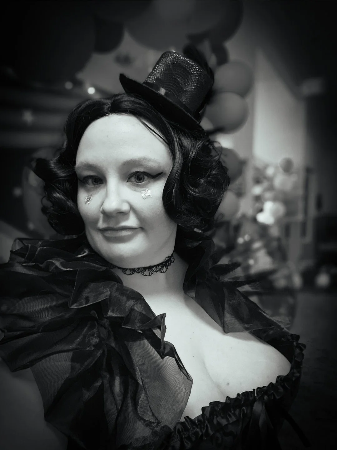 A woman with black curly hair and makeup featuring winged eyeliner and star-shaped decorations near her eyes, wearing a small black hat with some mesh detail, a black lace choker, and a ruffled black costume.
