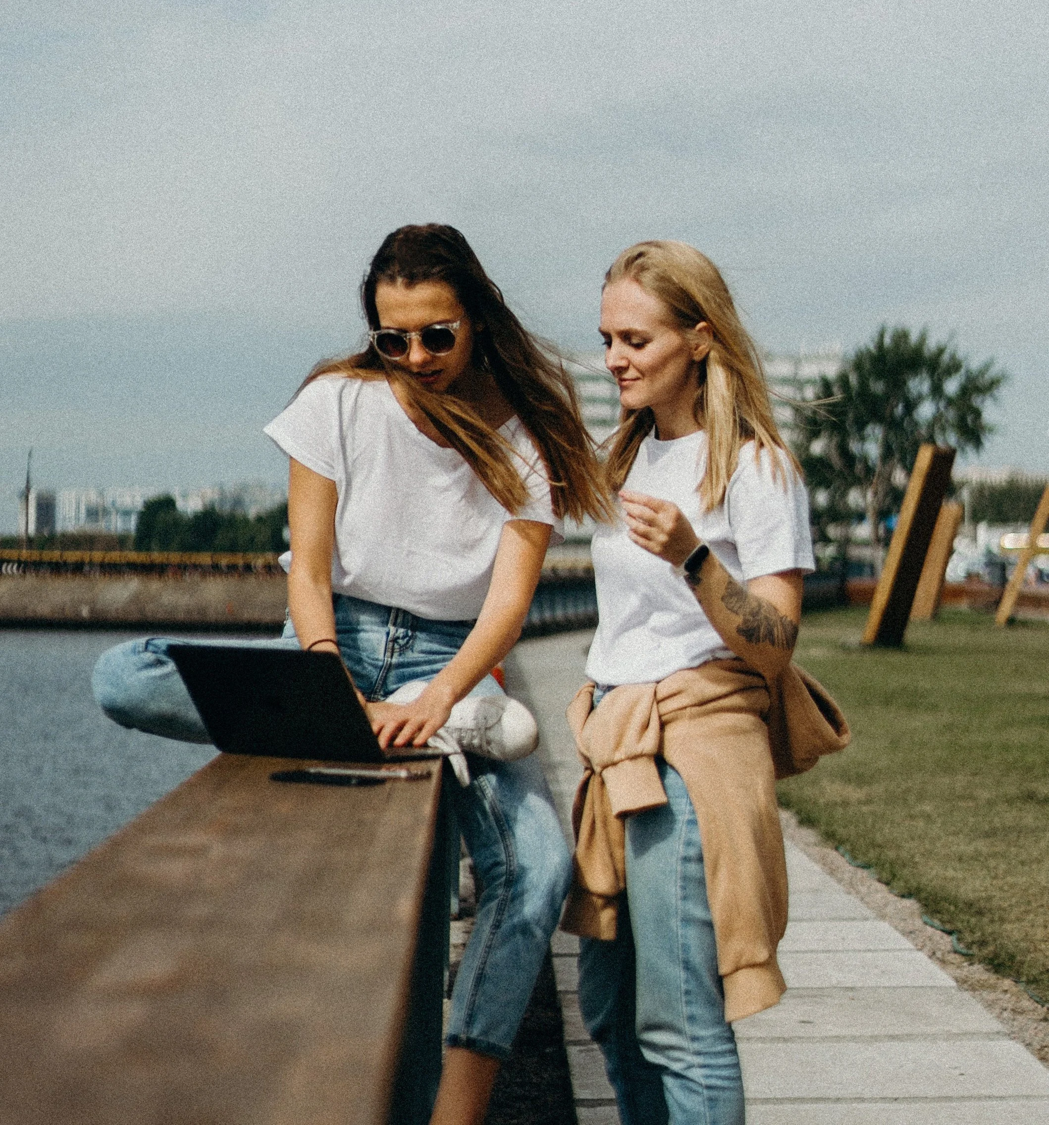 Two young women standing outdoors near a body of water, one wearing sunglasses and a white t-shirt, the other with long blonde hair and a tan coat draped over her arm, engaging in conversation on a cloudy day.