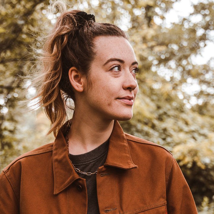 A young woman with curly hair tied up, wearing a brown jacket and a chain necklace, outdoors with blurred trees in the background.