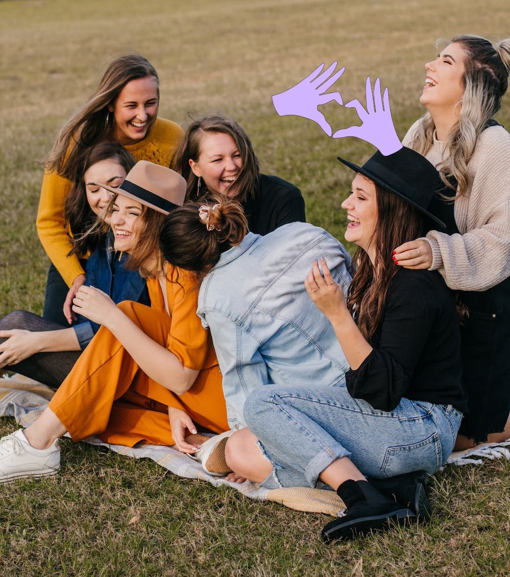 A group of seven young women sitting and lying on a blanket on the grass, smiling and laughing together outdoors.
