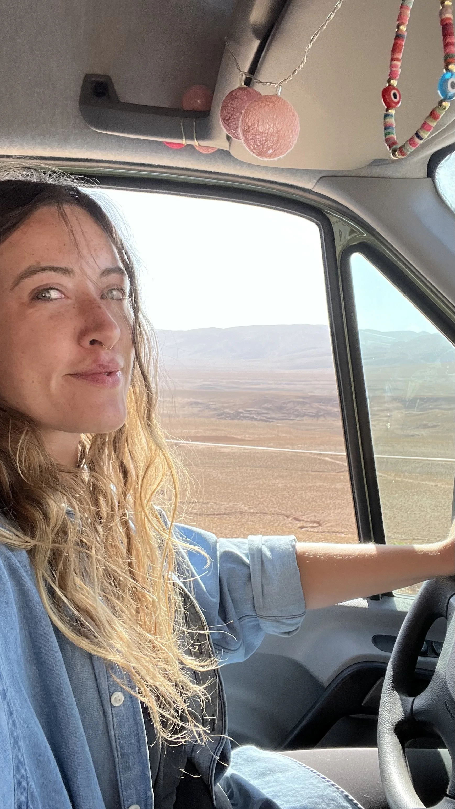 A woman with long, wavy blonde hair and a nose ring driving a vehicle in a desert landscape with mountains in the background. The interior has decorative hanging balls and a beaded necklace.