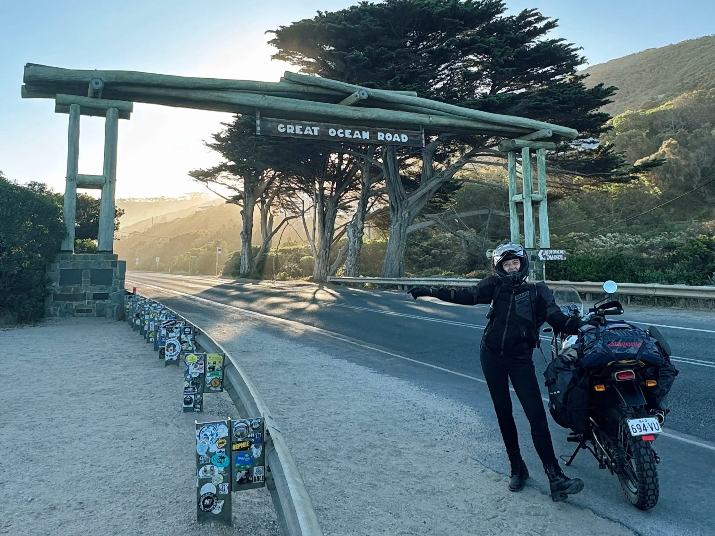 A person on a motorcycle standing next to a large sign that reads 'GREAT OCEAN ROAD' near the side of a highway. The sign is above a plaque with different stickers on it. The background features trees and hills, with the sun shining.