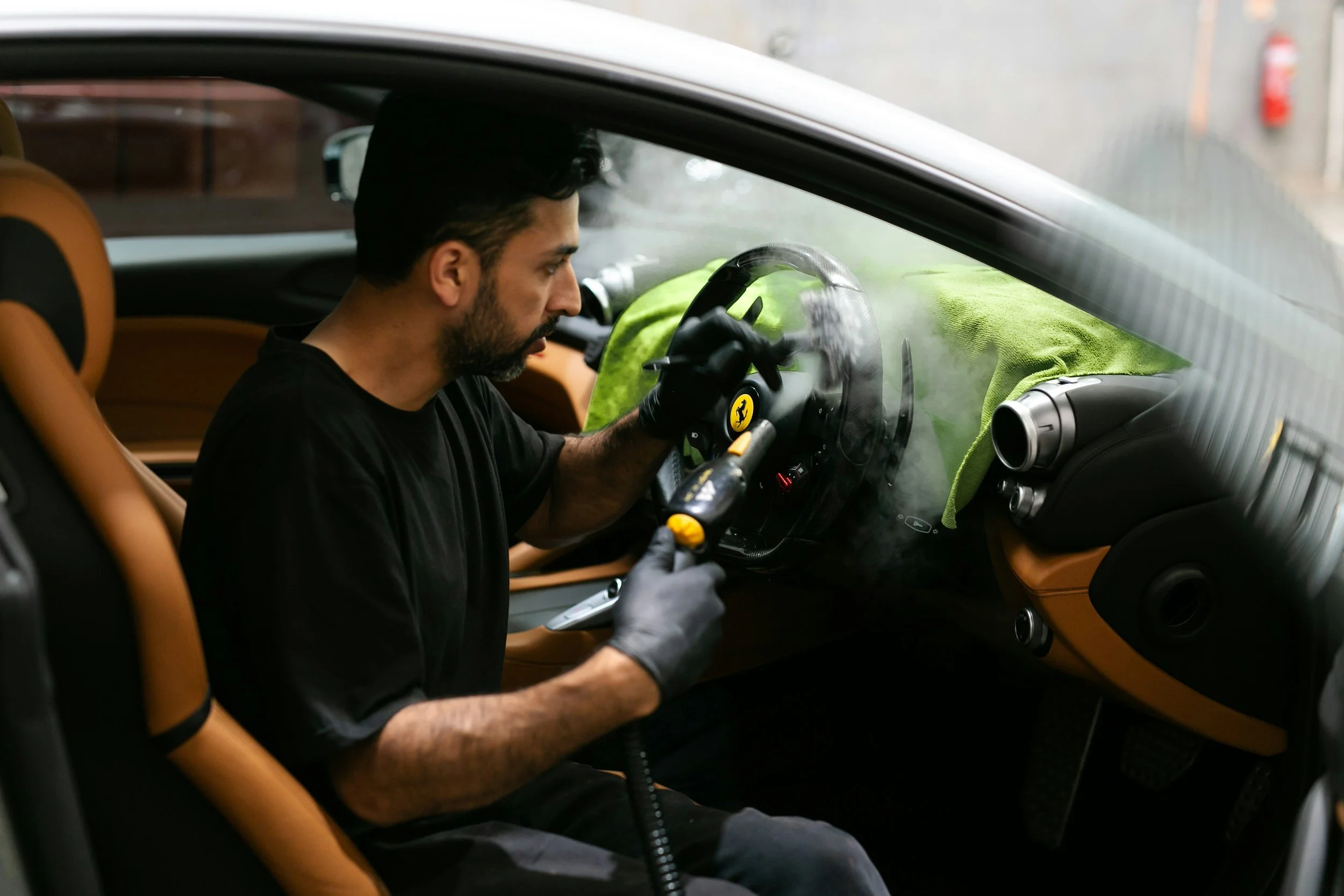 A man with dark hair and a beard, seated inside a luxury sports car, is holding a tool and working on the windshield, which has visible steam or smoke on it. The car's interior features tan leather seats and a black steering wheel with a Ferrari logo.