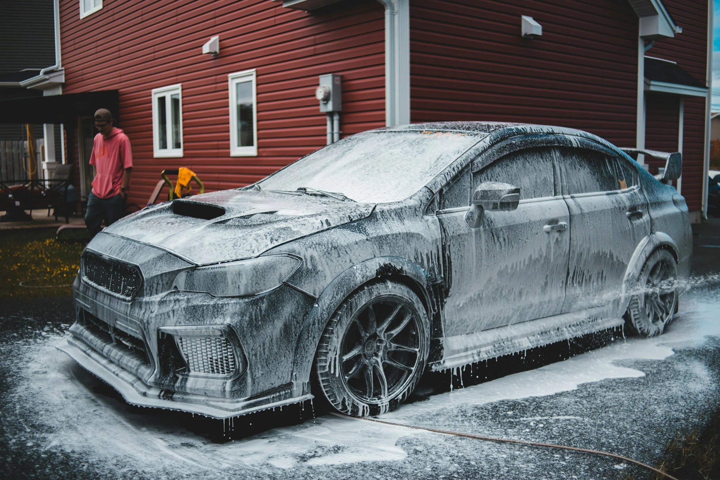 A black sports car is being washed with soap and foam, covered with white suds, in a driveway in front of a red house.