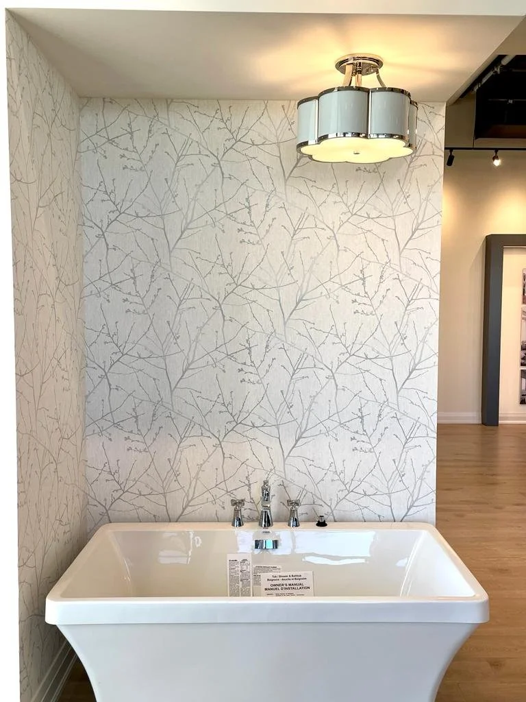 White bathtub with a faucet and two handles, placed against a wall with leaf-patterned wallpaper. There is a modern ceiling light fixture above the tub and a partial view of a room with wooden flooring and a display frame in the background.
