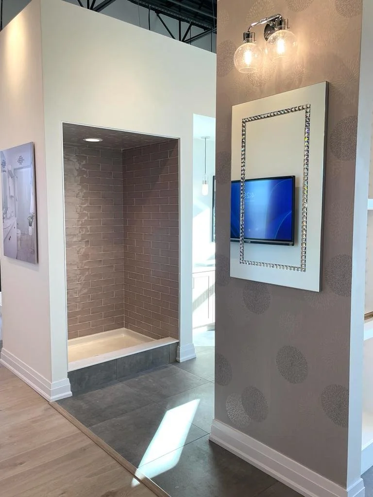 An interior space with a walk-in shower featuring brown brick tiles, with a modern flat-screen television mounted on a wall with decorative wallpaper and a mirror framed with rhinestones; hanging above is a light fixture with two glass globes.