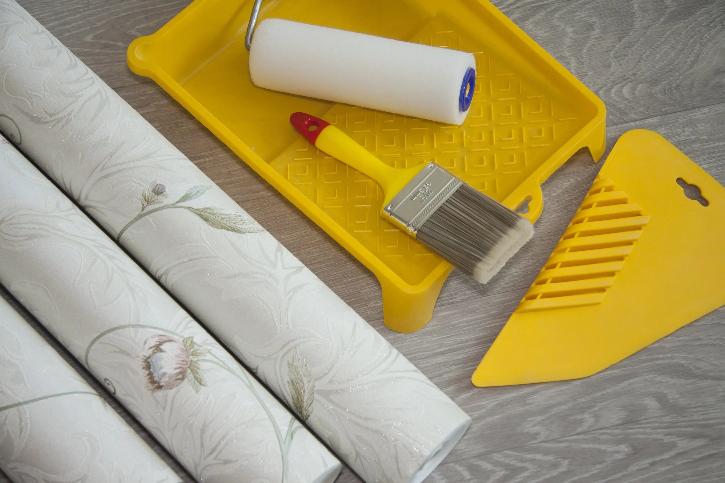 Wallpaper rolls, paint roller with white paint, yellow paint tray, brush, and spatula placed on a light-colored wooden floor.