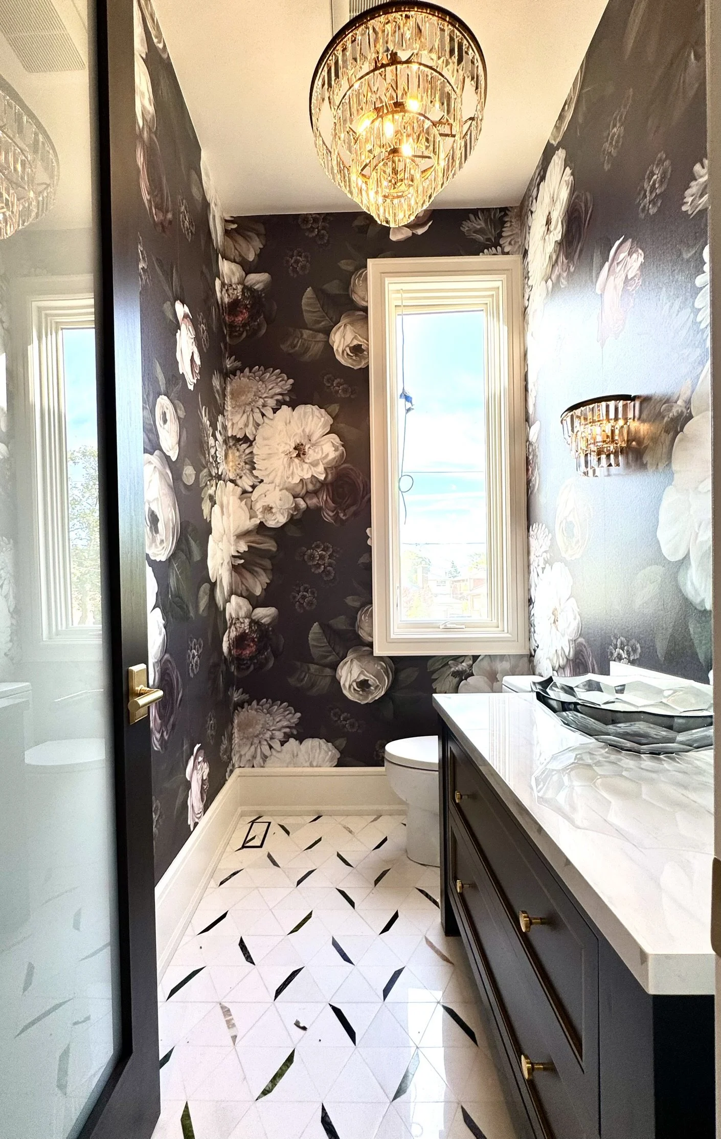 A bathroom with black floral wallpaper by Ellie Cashman, a window with a view of the sky, a chandelier, wall sconce, a white toilet, and a dark vanity with a white marble countertop.