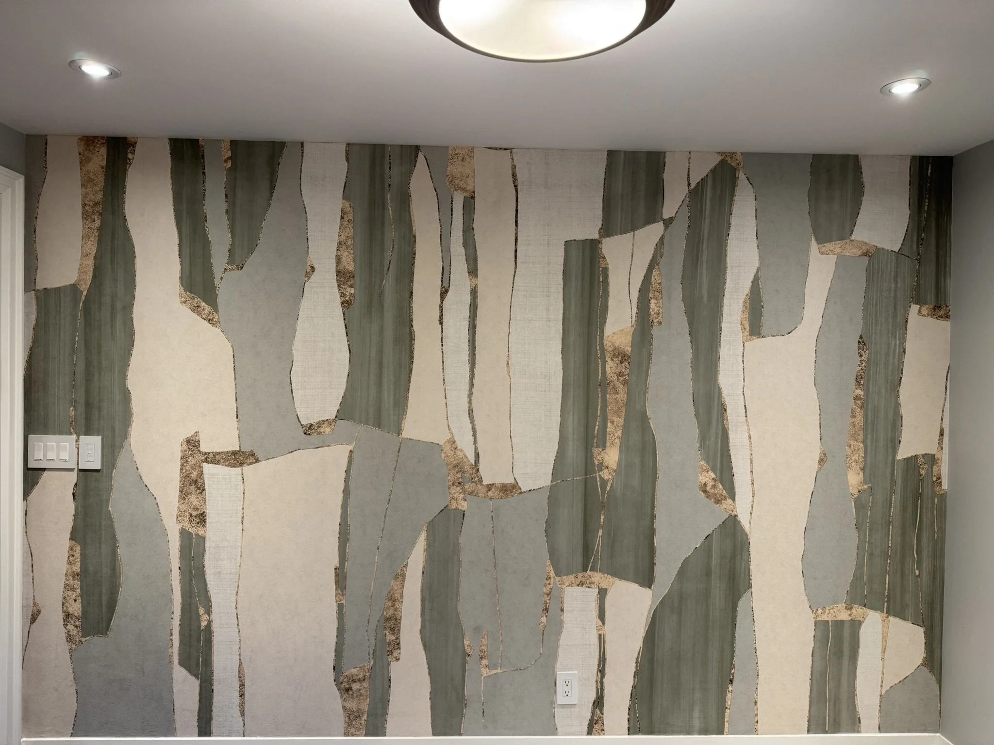 Living room wall with abstract patterned wallpaper by Tecnografica featuring irregular green, beige, and brown shapes. Recessed ceiling lights and electrical switches are visible.
