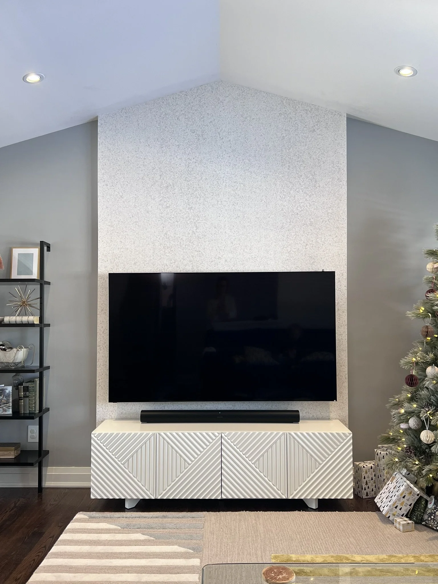 Living room with a large flat-screen TV mounted on a textured wallpaper by Phillip Jeffries, white TV stand beneath, a decorated Christmas tree on the right side, and a black shelf on the left with various decor items. 