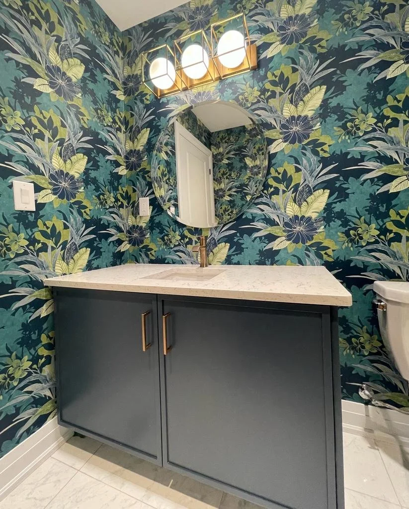 A bathroom with dark blue floral wallpaper, a round mirror, a white countertop with an integrated sink, a dark blue cabinet with gold handles, a modern light fixture with three globe bulbs, and a white toilet partially visible on the right.