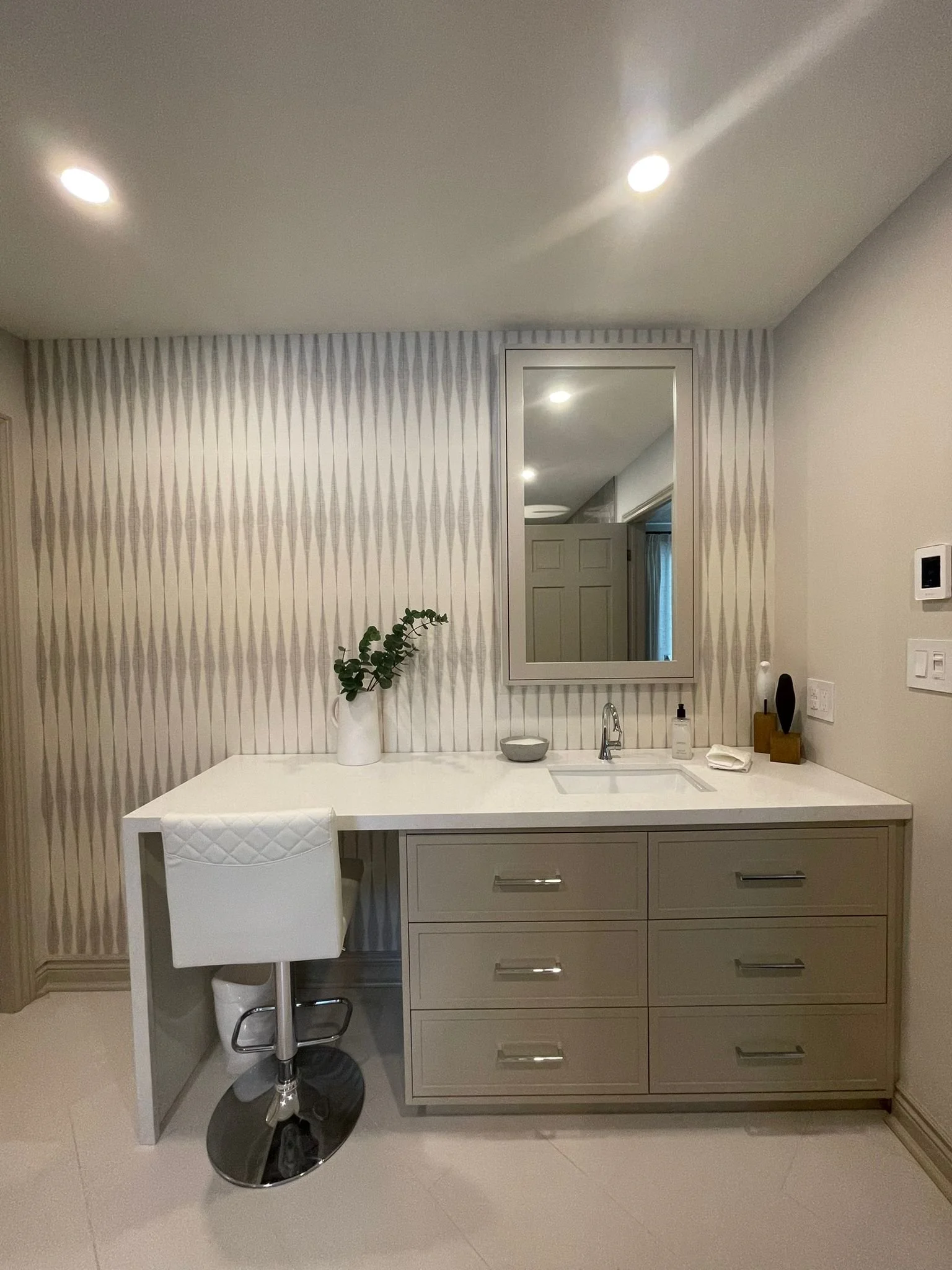 A modern bathroom vanity with a white countertop, gray drawers, a small sink, a mirror above, a white chair with quilted upholstery in front, and decorative items including a vase with greenery, a soap dispenser, a bowl, and small decorative objects.