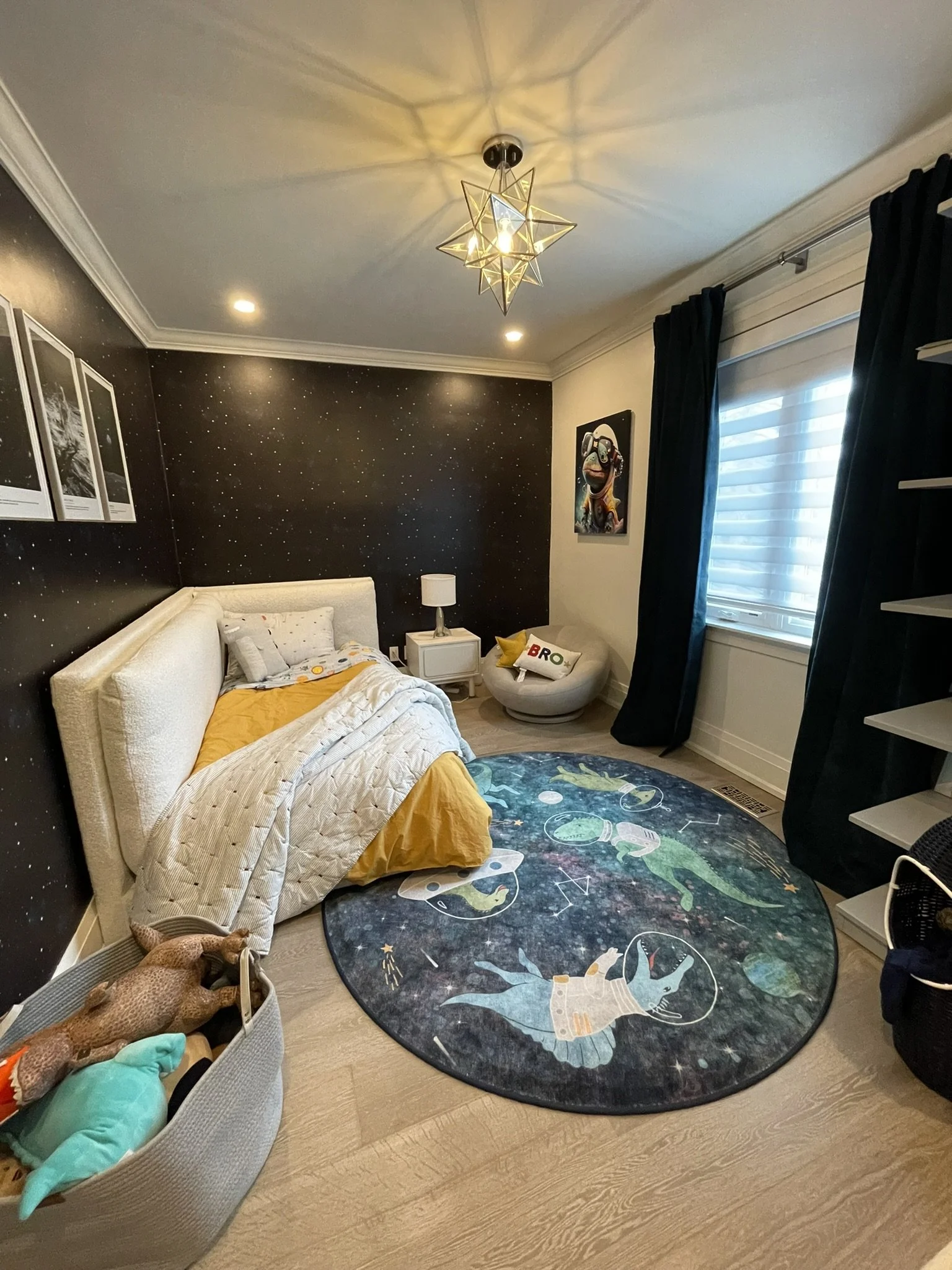 A bedroom with a space-themed decor. It has a black starry wallpaper by Spoonflower, a white bed with yellow bedding, a round rug with space and astronaut designs.