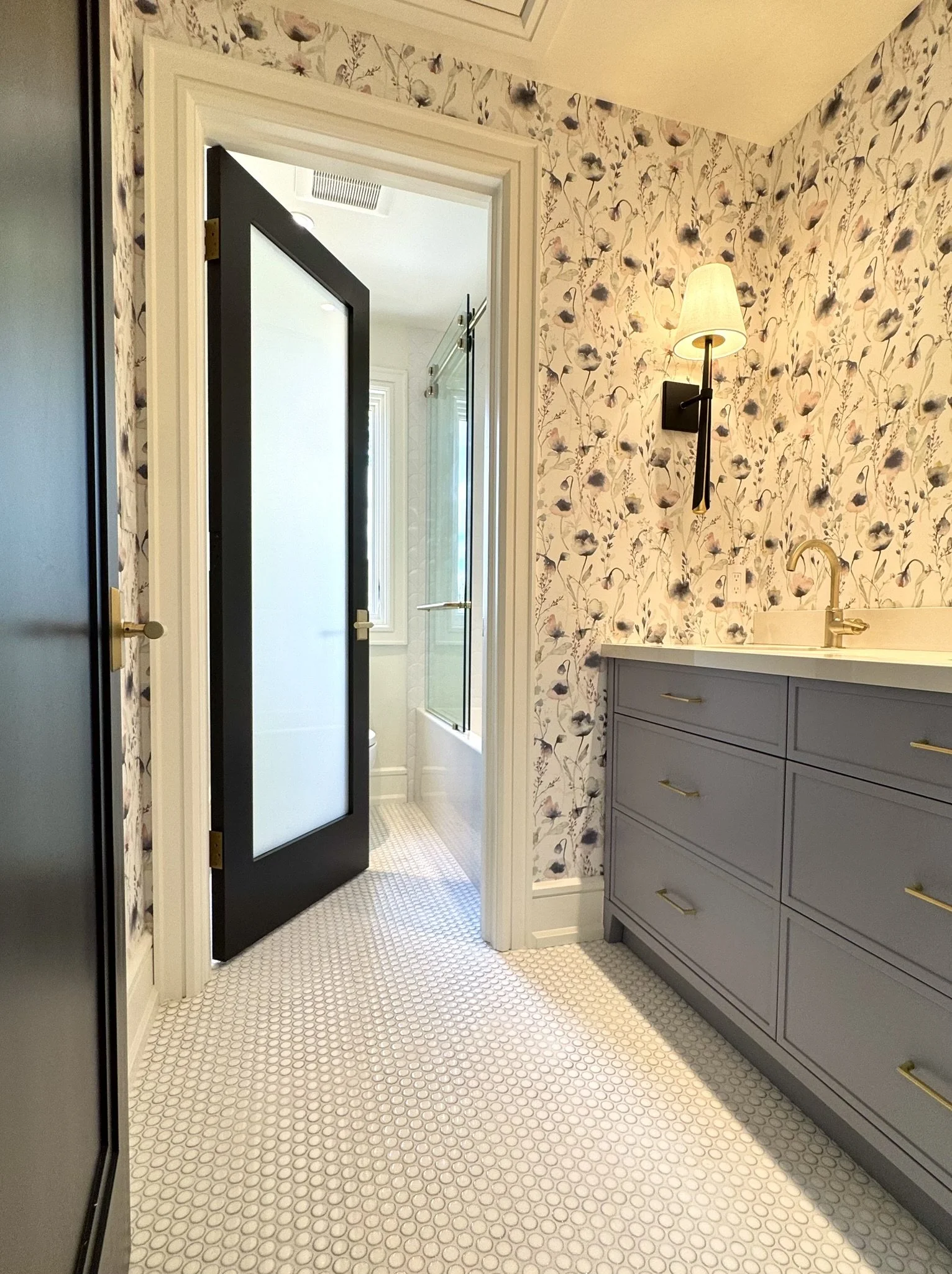 A bathroom with floral wallpaper by Sandberg Wallpaper , a black wall sconce with a white lampshade, a gray vanity with gold handles, a white countertop, a gold faucet, and a black door.