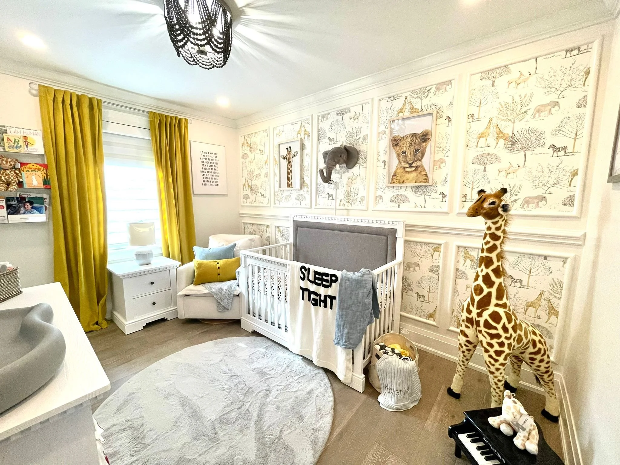 Nursery room with yellow curtains, wall art featuring animals, a white crib, a stuffed giraffe, a comfortable armchair with pillows, a window with blinds, and a toy piano. A sign on the crib reads 'Sleep Tight'.