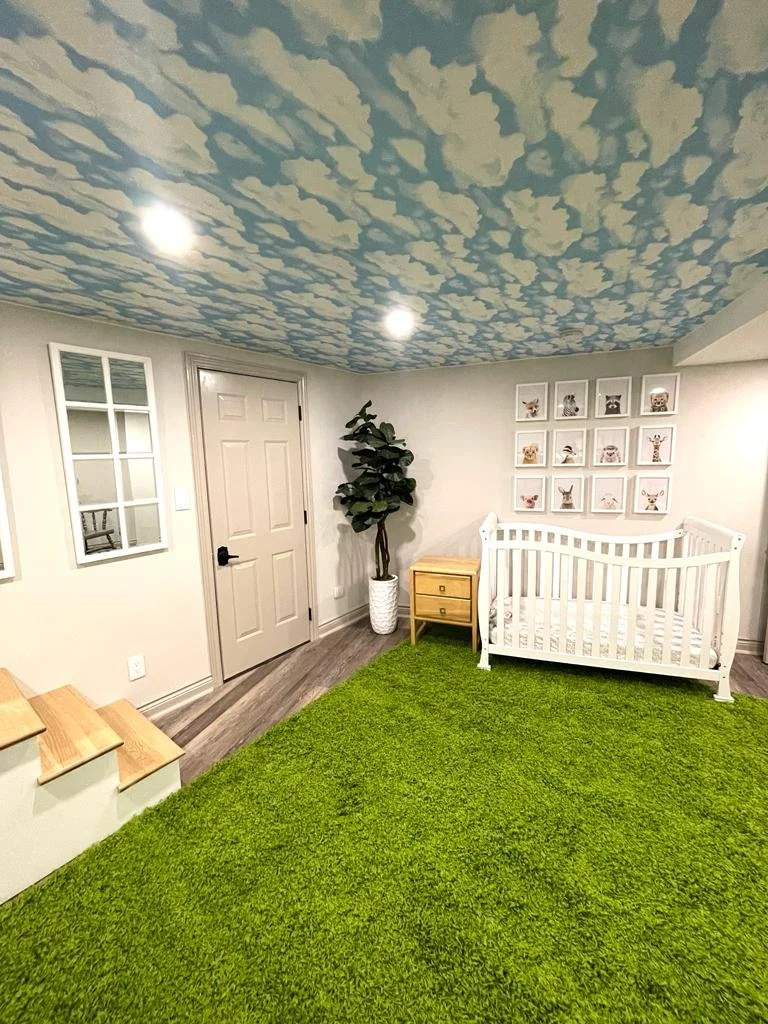A room with a sky ceiling decorated with clouds, a white crib, a small wooden chest of drawers, a tall plant in a white pot, framed animal pictures on the wall, a green rug, and wooden steps.