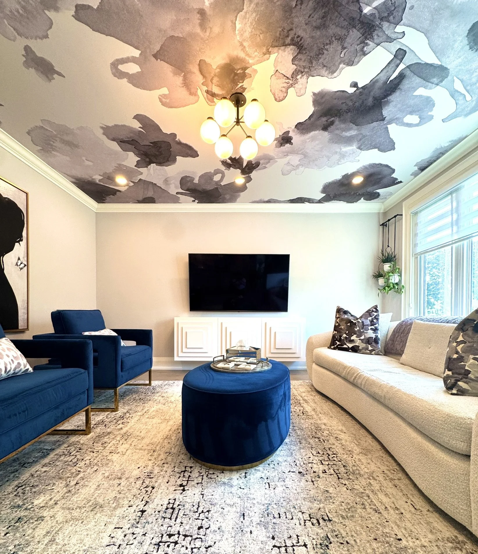 Living room with a colorful abstract ceiling mounted wallpaper by Drop it Modern , navy blue chairs, a beige sofa, and a matching navy blue ottoman in the centre.