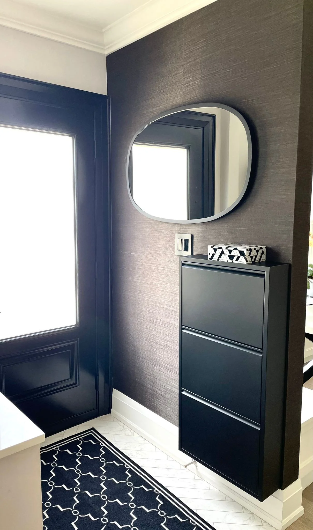 Corner of a modern interior with black cabinet, a round mirror, a geometric patterned tissue box, white trim, and a patterned black and white rug on white flooring.