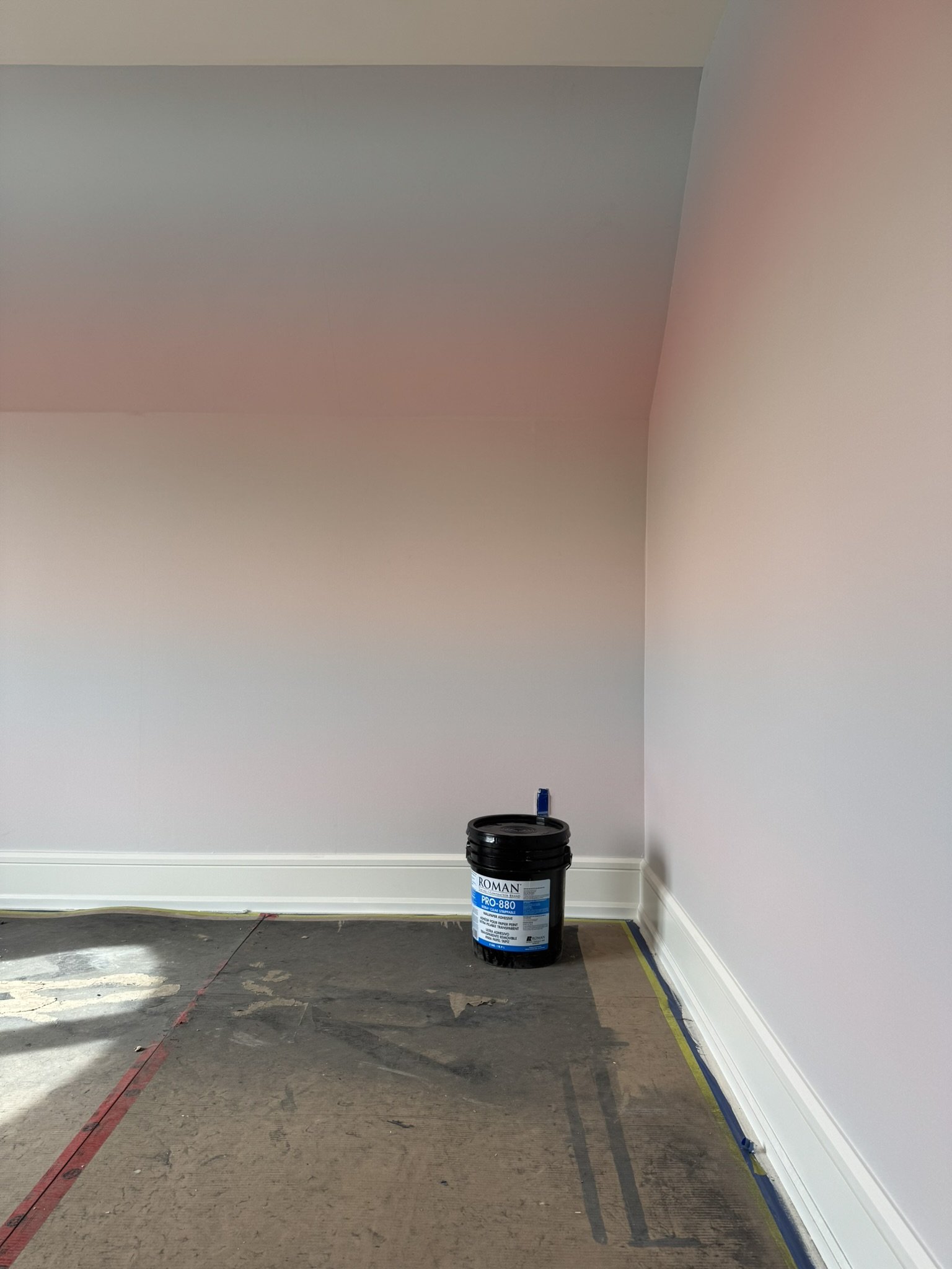 Corner of a room with wallpaper by Drop It Modern, with a white baseboard, brown floor, and a black bucket labeled 'Roman PRO-880' in the corner.