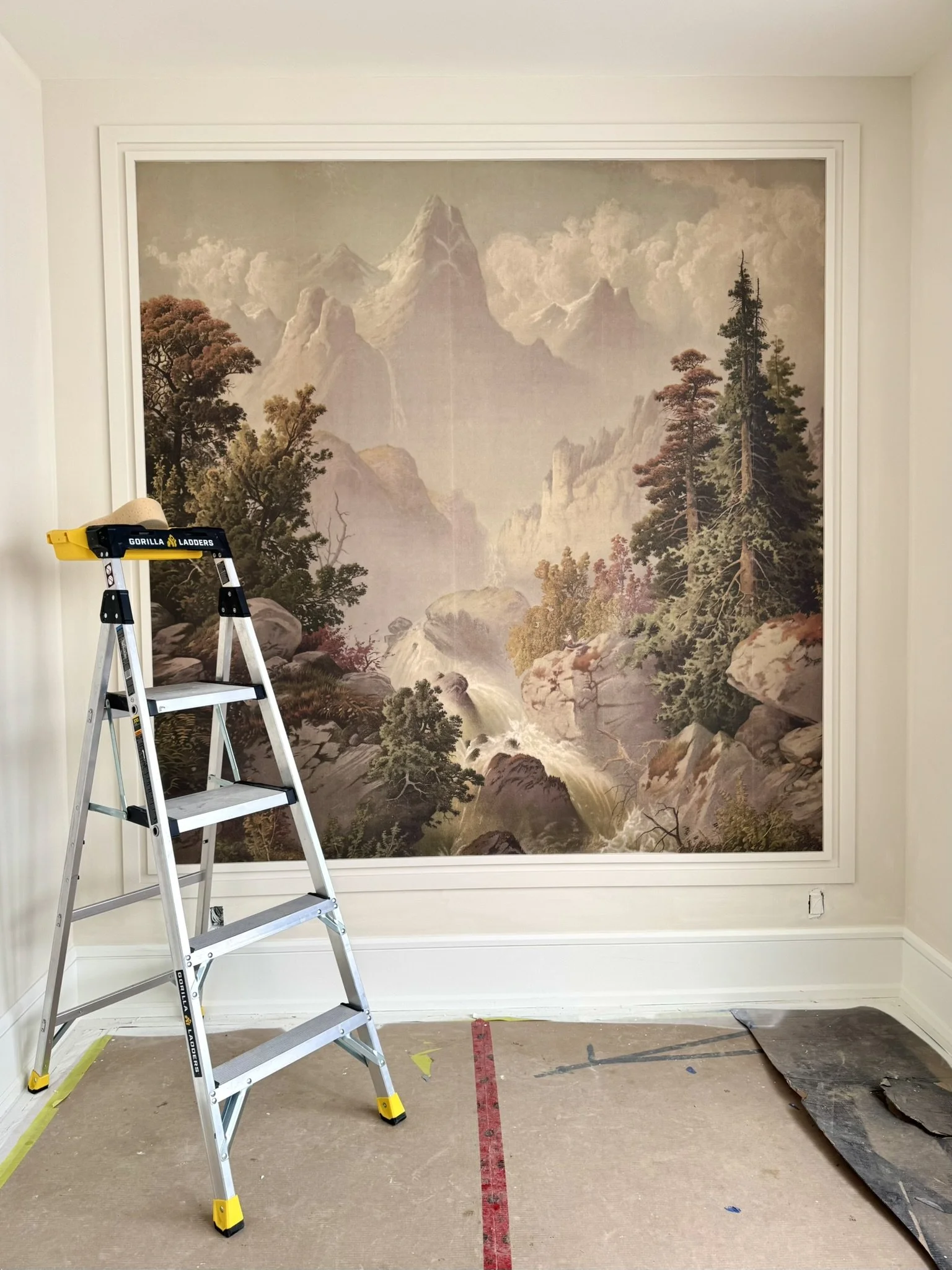 A large wallpaper mural of a mountain landscape with trees by Rebel Walls, rocks, and a waterfall, designed to look like a painting. 