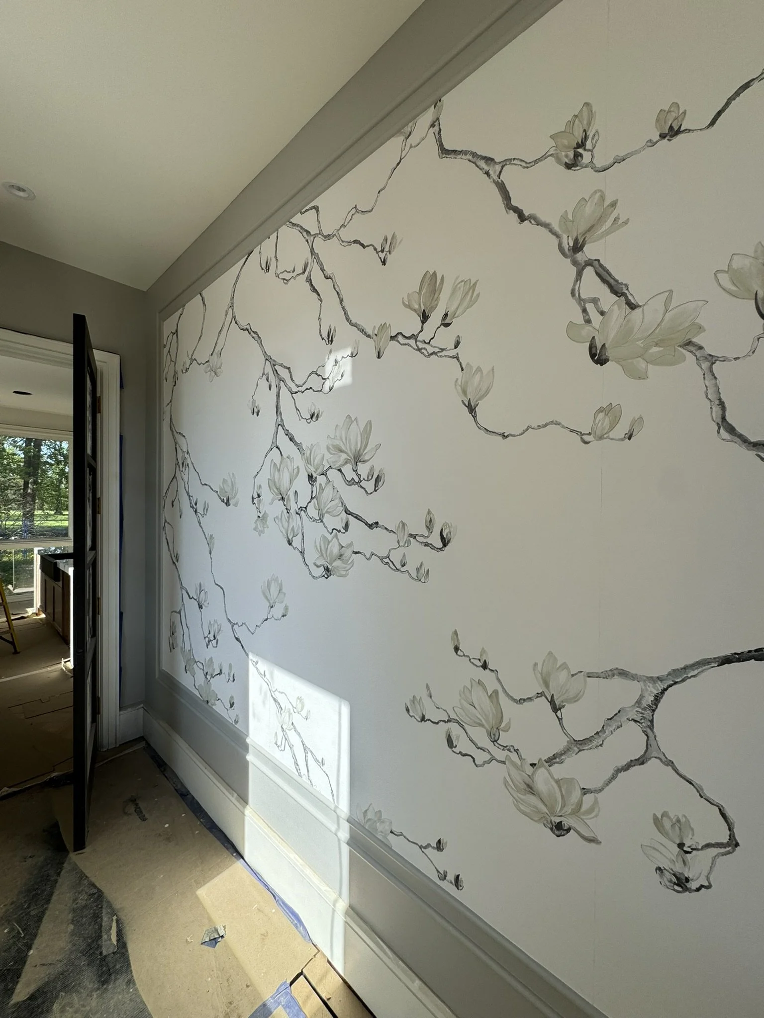 Interior wall with floral mural by Phillip Jeffries featuring branches and white flowers, near an open door leading to a room with a window.