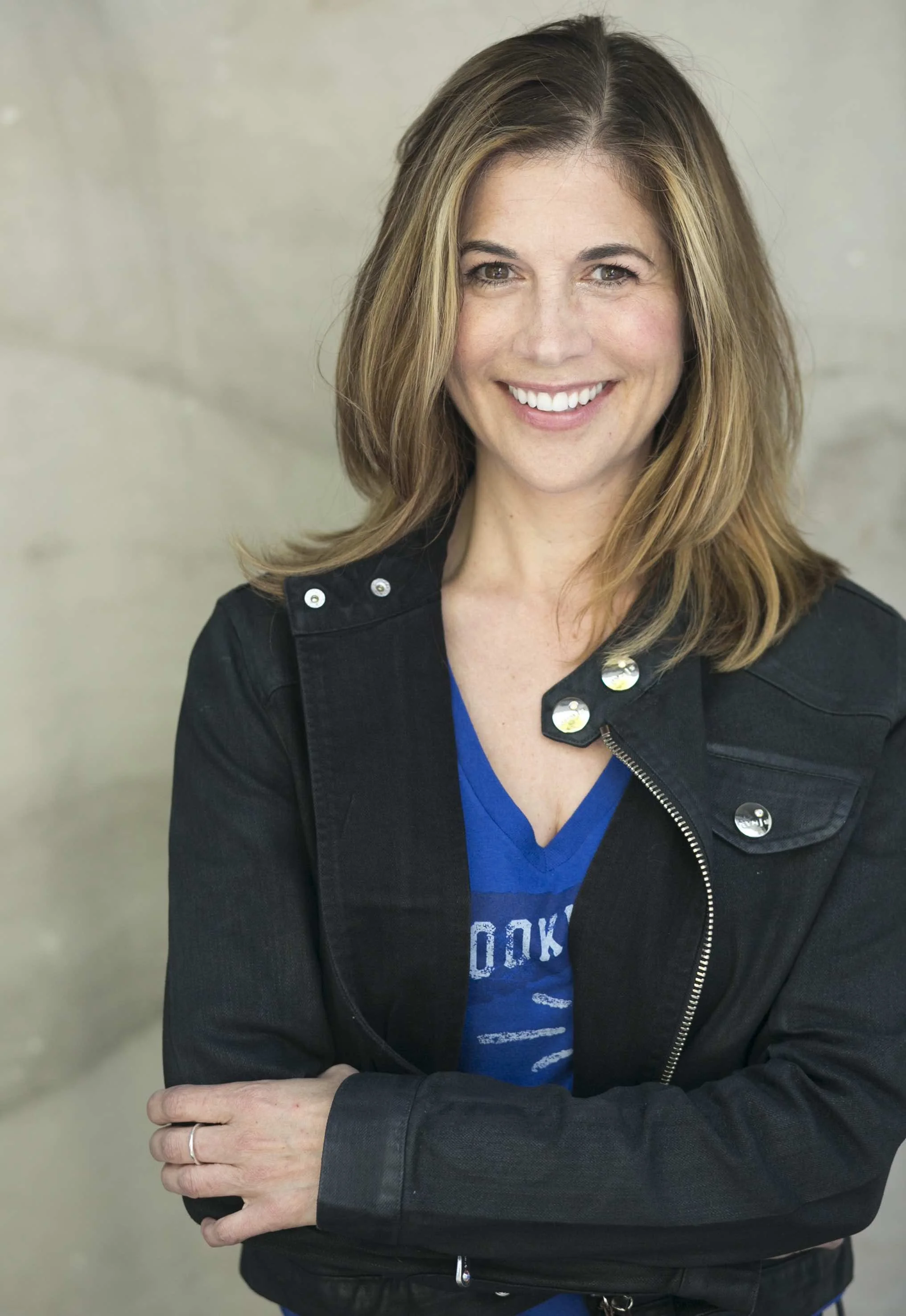 A woman with shoulder-length light brown hair smiling, wearing a black jacket over a blue shirt, standing with arms crossed in front of a neutral background.