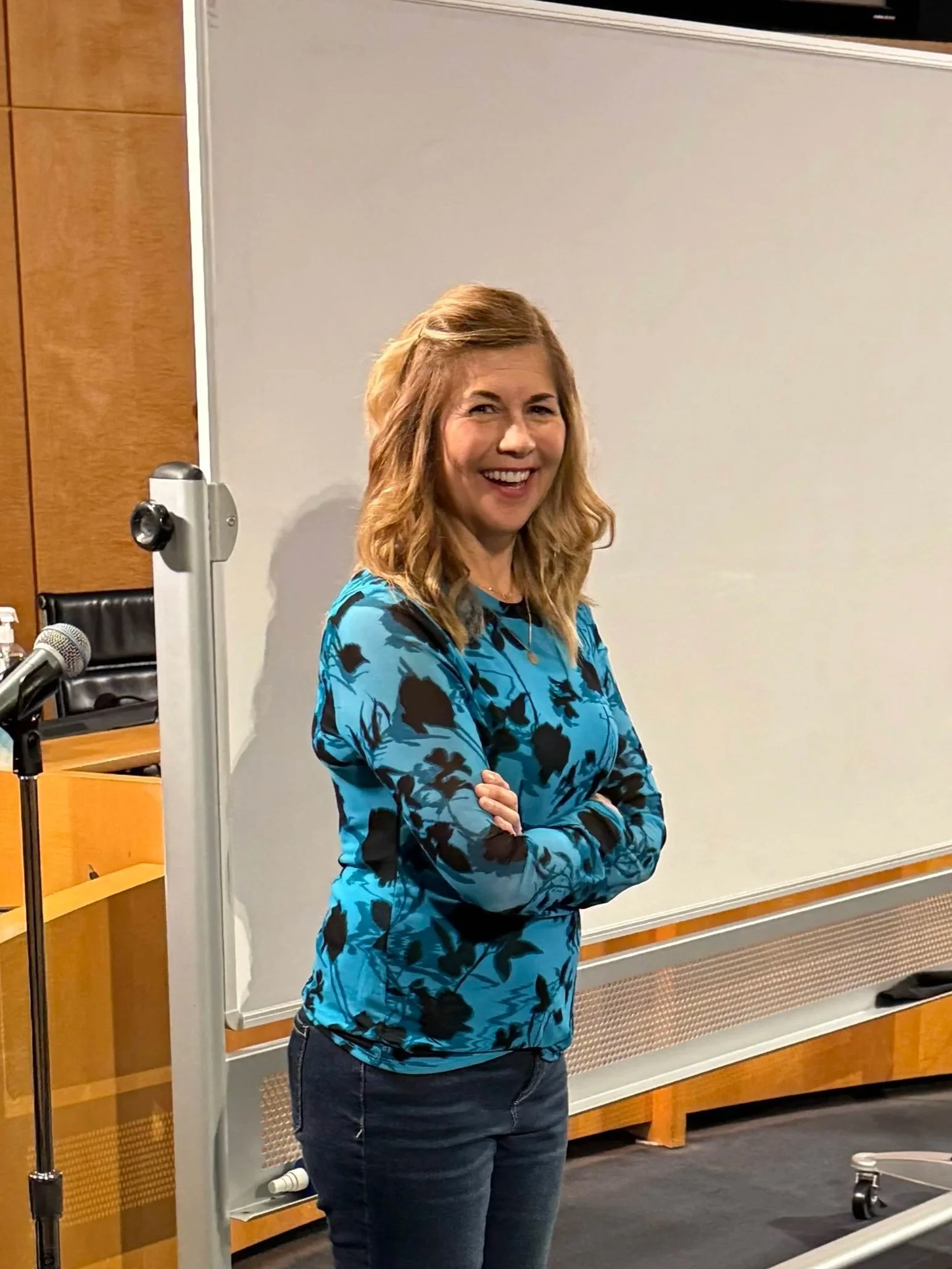 A woman with shoulder-length red hair, smiling, standing with crossed arms in front of a whiteboard in a conference room, wearing a blue floral top and dark jeans.