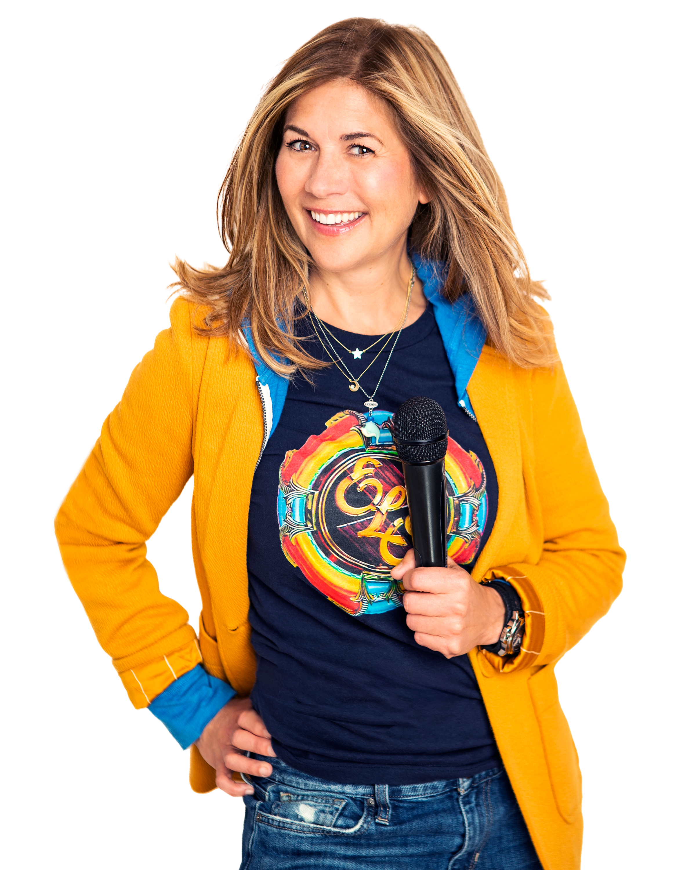 A woman smiling while holding a microphone, wearing a yellow jacket over a graphic T-shirt, with layered necklaces and a watch.