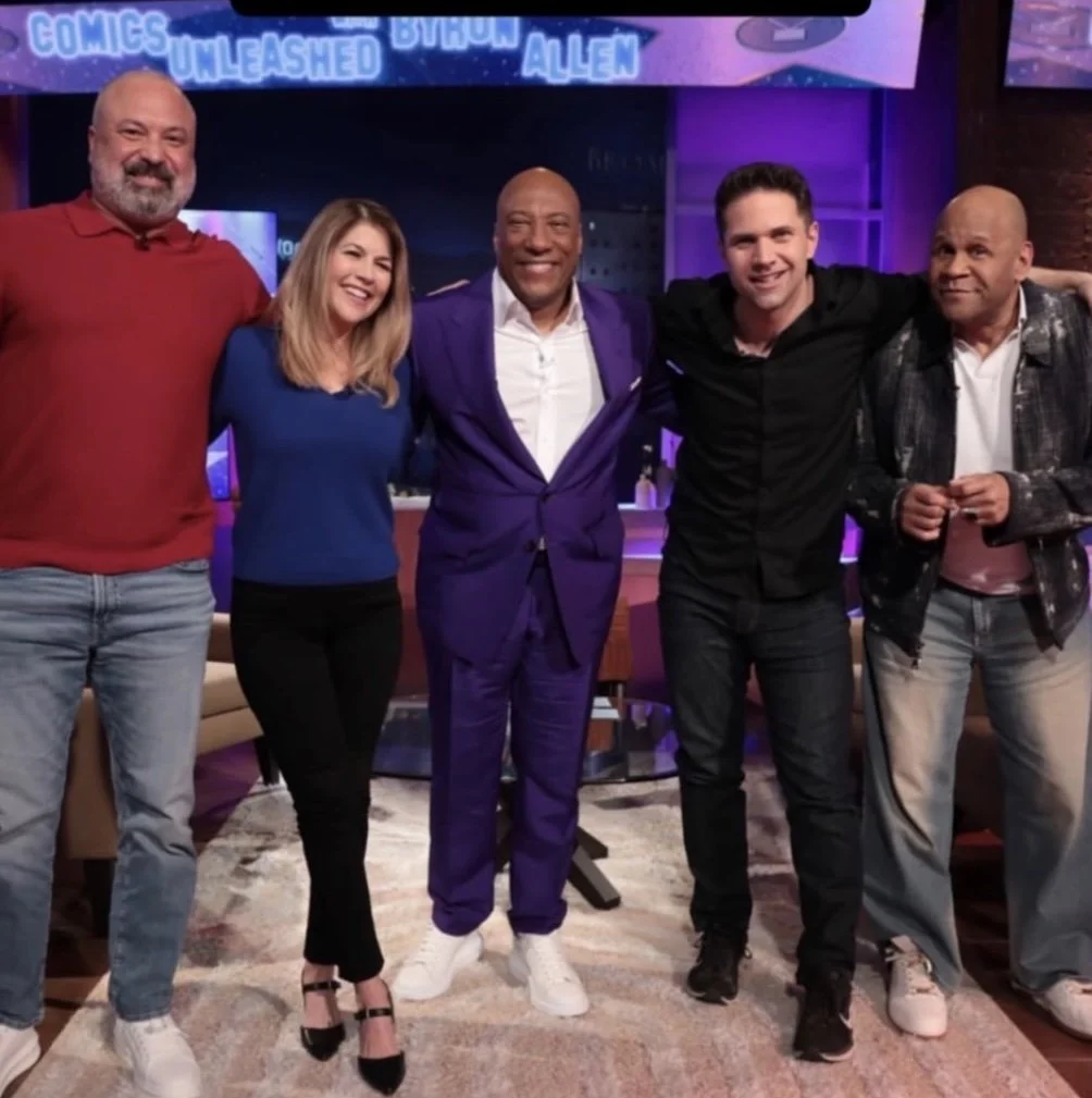 Five comedians posing for a picture with arms on each others shoulders. Mary is the second from left in a blue shirt and black pants.