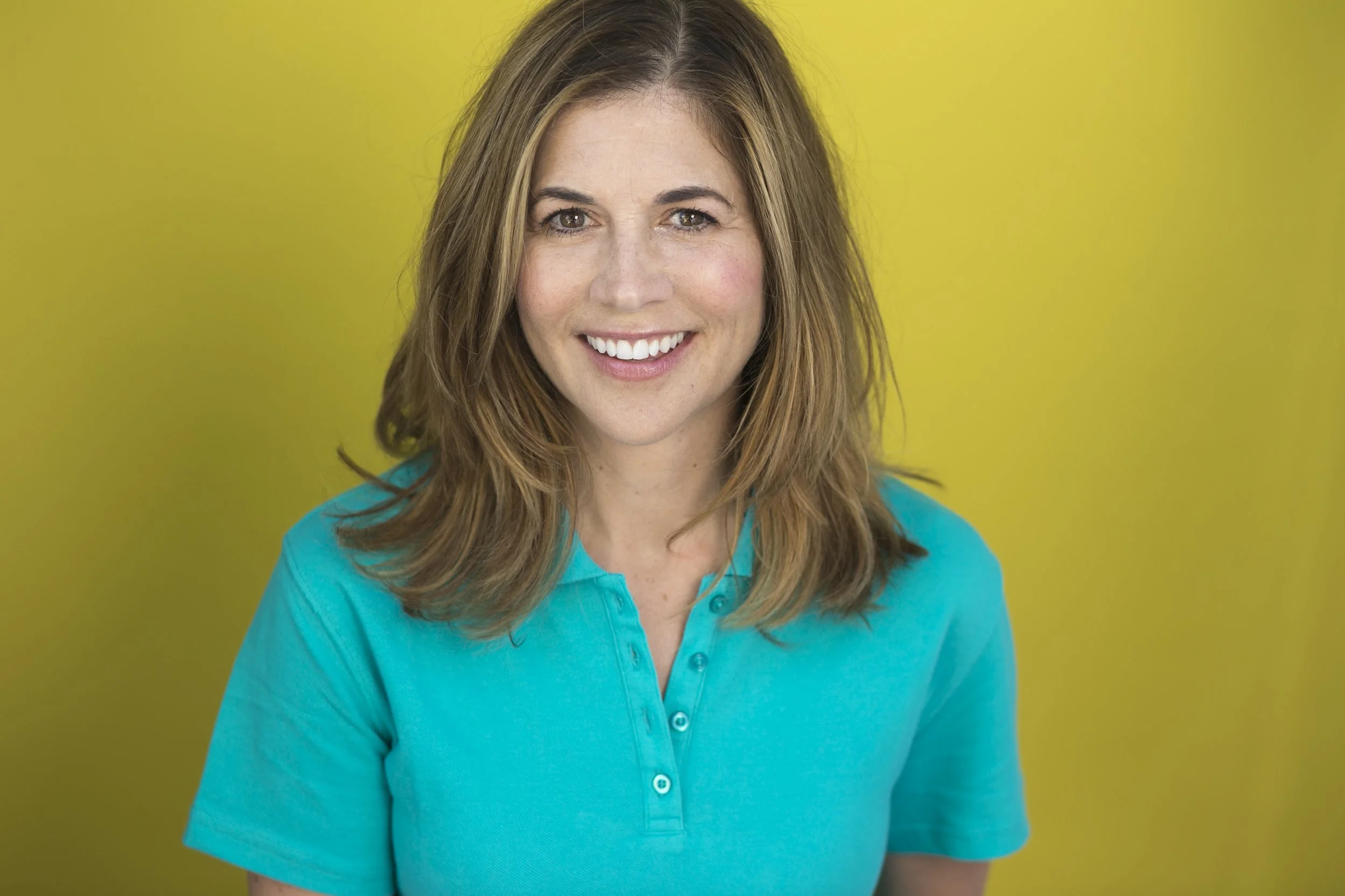 A woman with shoulder-length light brown hair wearing a teal top smiling in front of a yellow background.